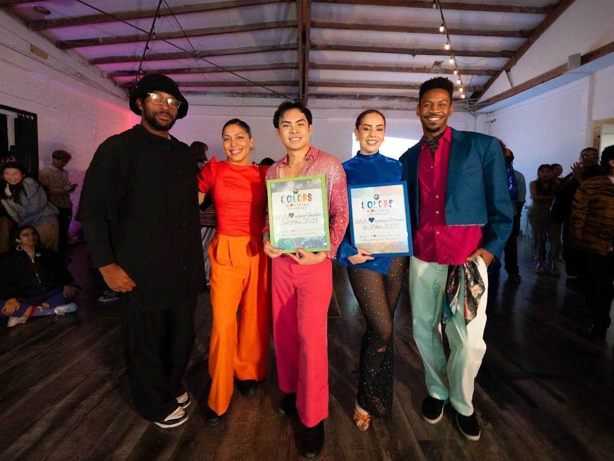 Group of five people at an event, two holding colorful framed certificates, posing and smiling in a brightly lit venue with exposed wooden ceiling and other attendees in the background.