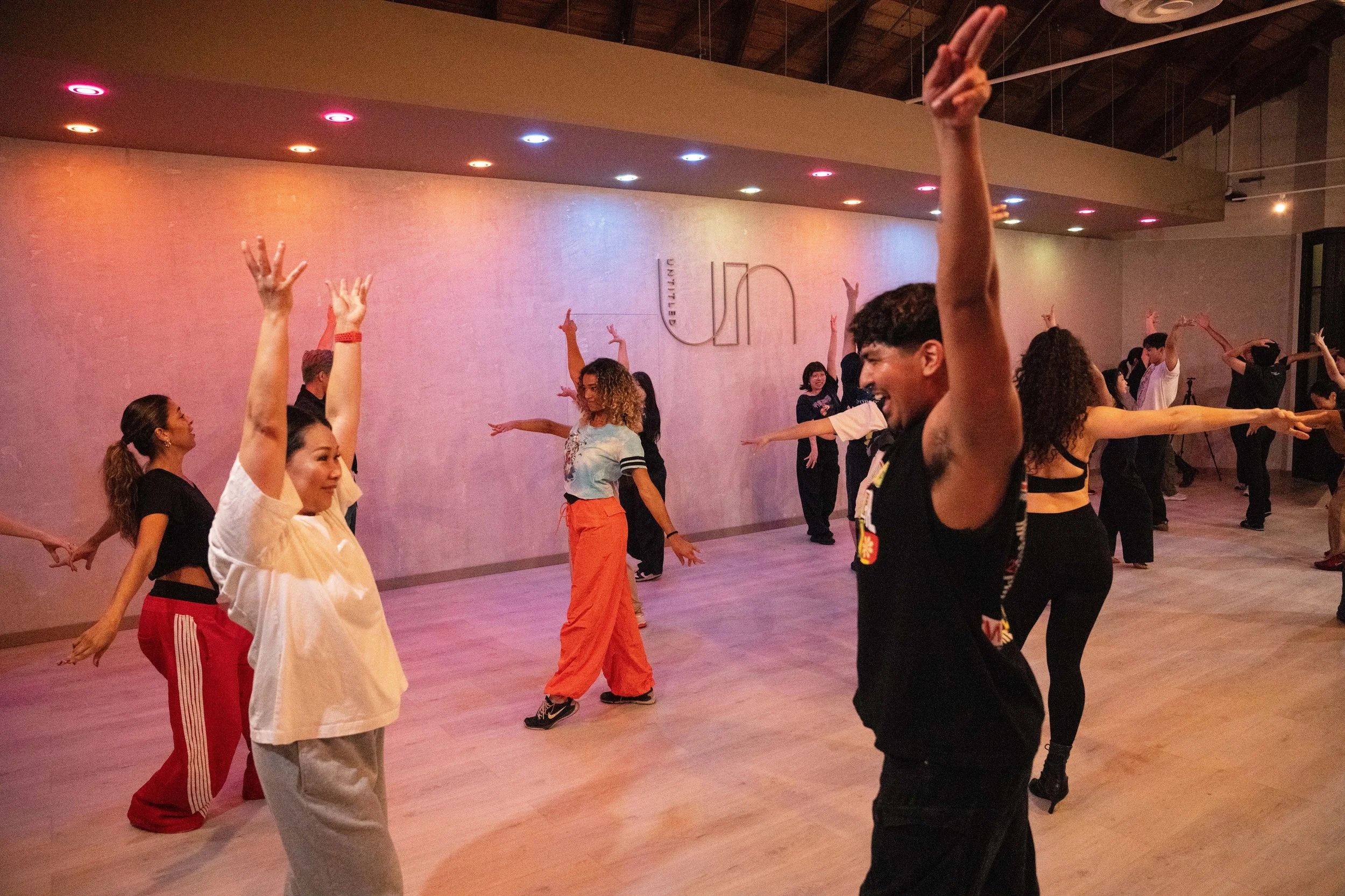 Group of diverse people dancing in a studio with wooden floors, in a synchronized dance class or rehearsal, illuminated by colored ceiling lights.
