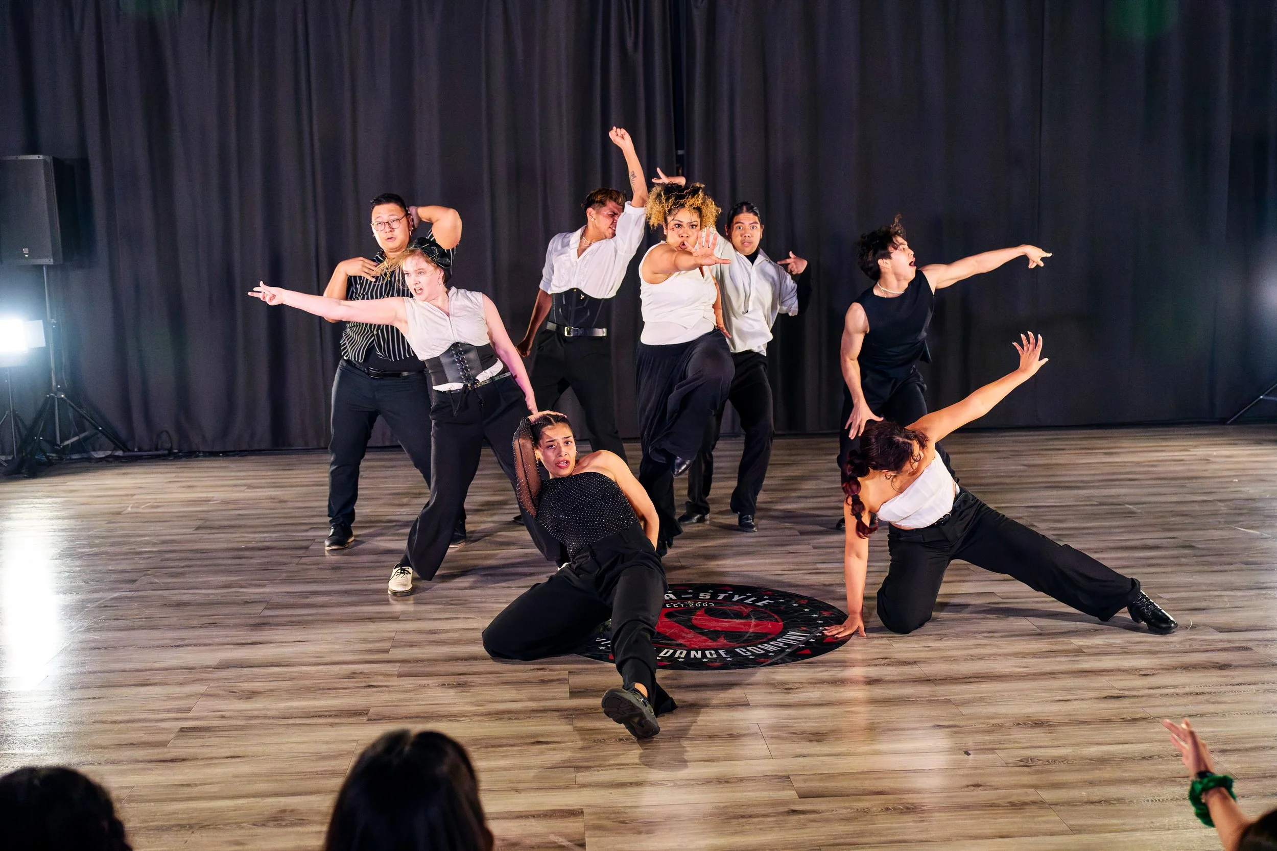 A group of diverse dancers performing on stage with black curtains in the background. They are striking various expressive poses and wearing stylish dance costumes.