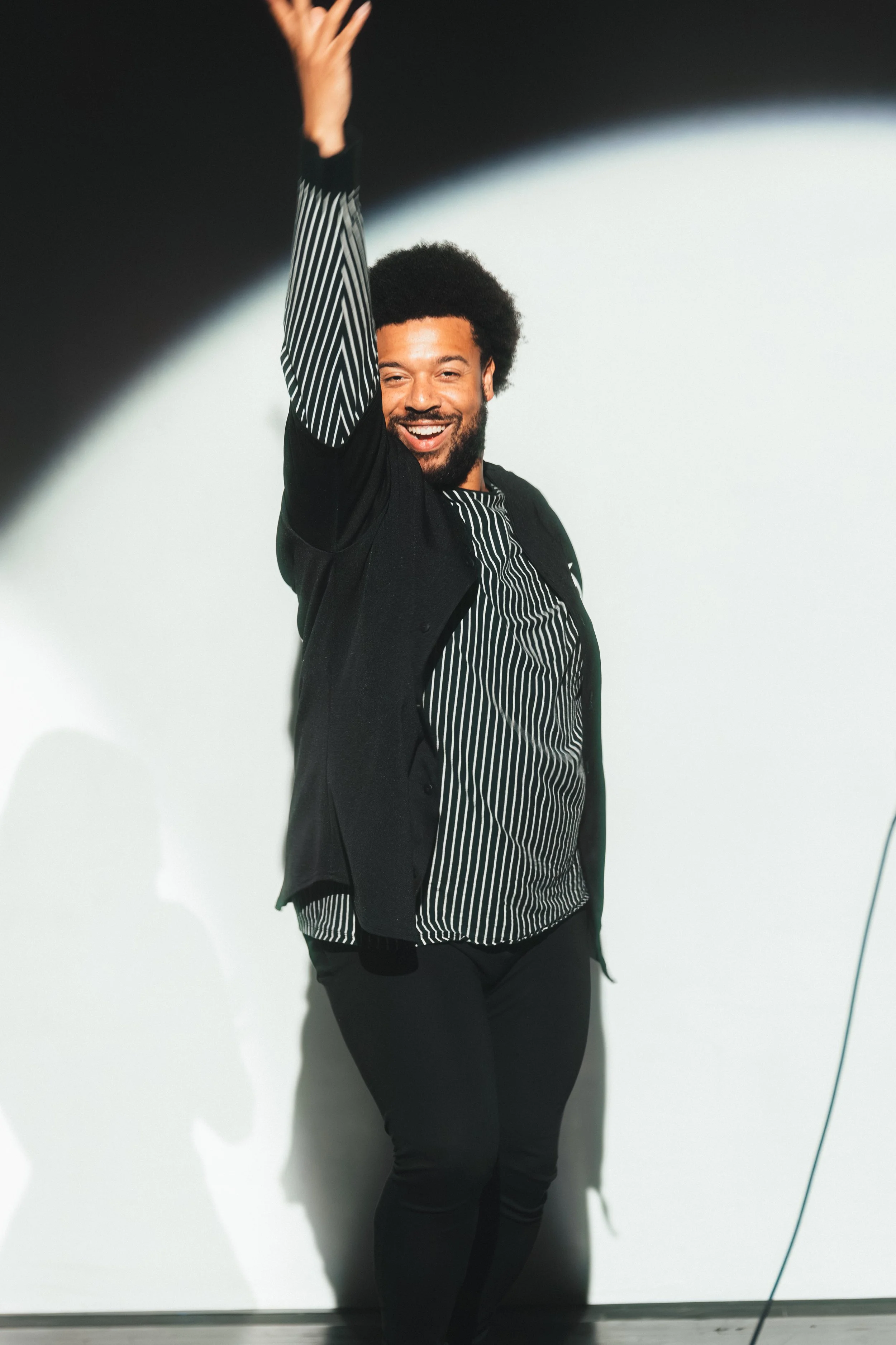A man with a beard and afro hairstyle smiling and raising his right hand in a black and white striped shirt with a black jacket, standing against a plain light background.