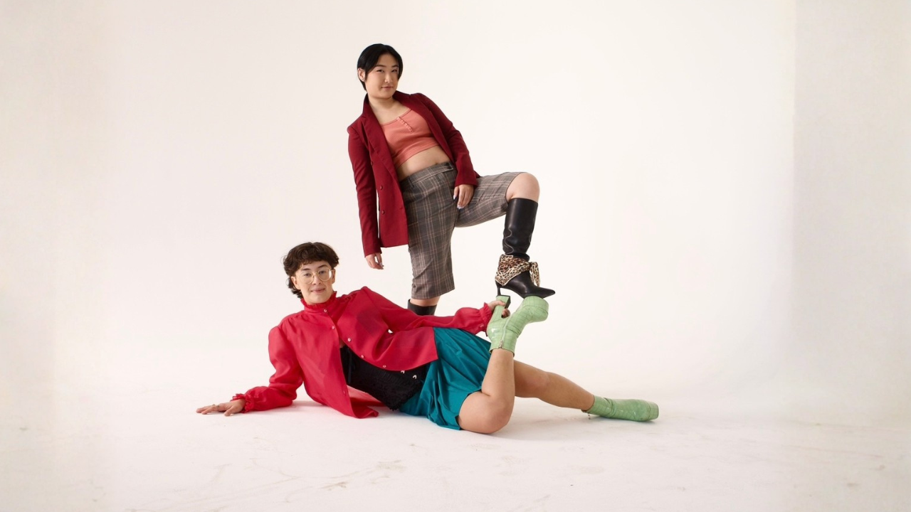 Two women posing in a studio with a plain white background. One is lying on the floor with one arm supporting her, wearing a red jacket, teal shorts, green socks, and glasses. The other is standing on her colleague's knee with one foot, wearing a red