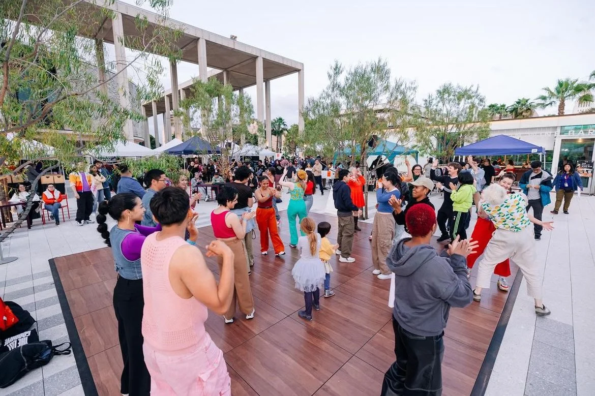 People dancing and socializing at an outdoor event with trees and tents in the background.