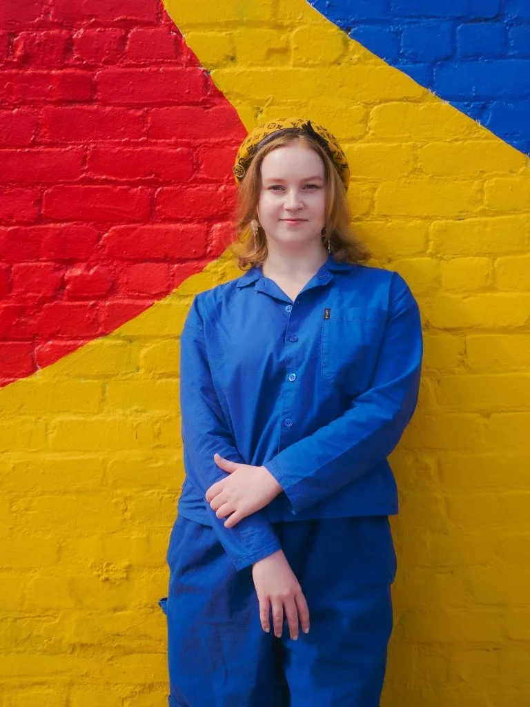 A young woman with shoulder-length blond hair, wearing a yellow bandana, a blue long-sleeve shirt, and matching blue pants, standing against a brightly painted brick wall with red, yellow, and blue sections.