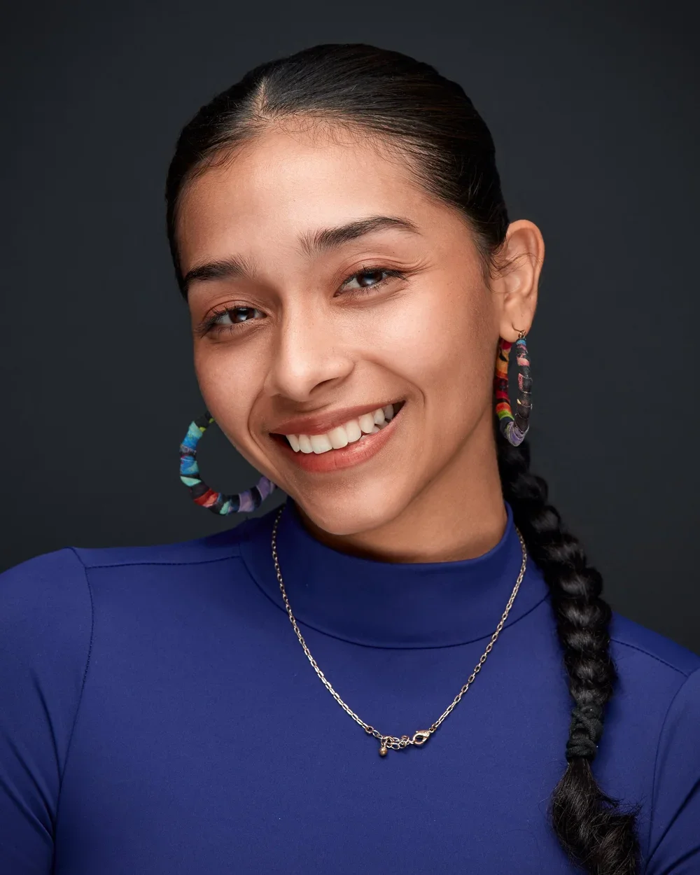 Portrait of a young woman with dark hair in a braid, wearing a blue top, colorful earrings, and a silver necklace, smiling against a dark background.