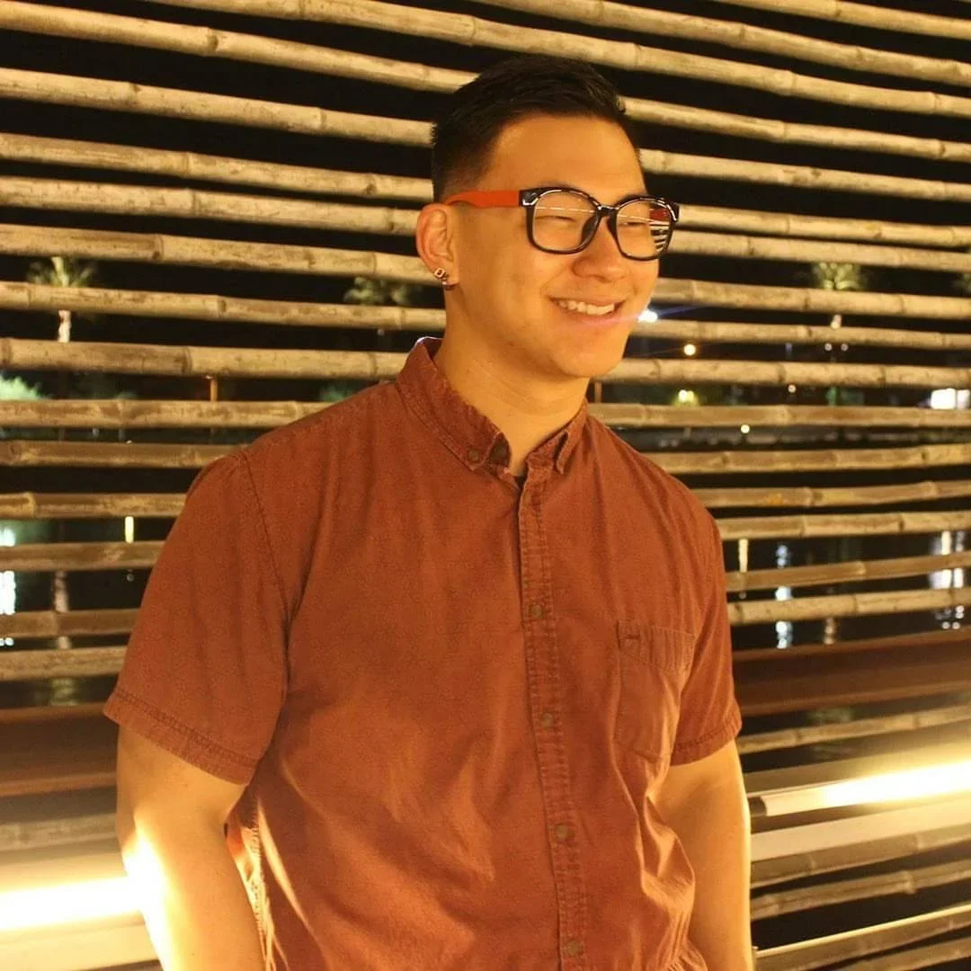 A young man wearing large glasses and a rust-colored button-up shirt, smiling with eyes closed, standing against a background of horizontal wooden slats at night.
