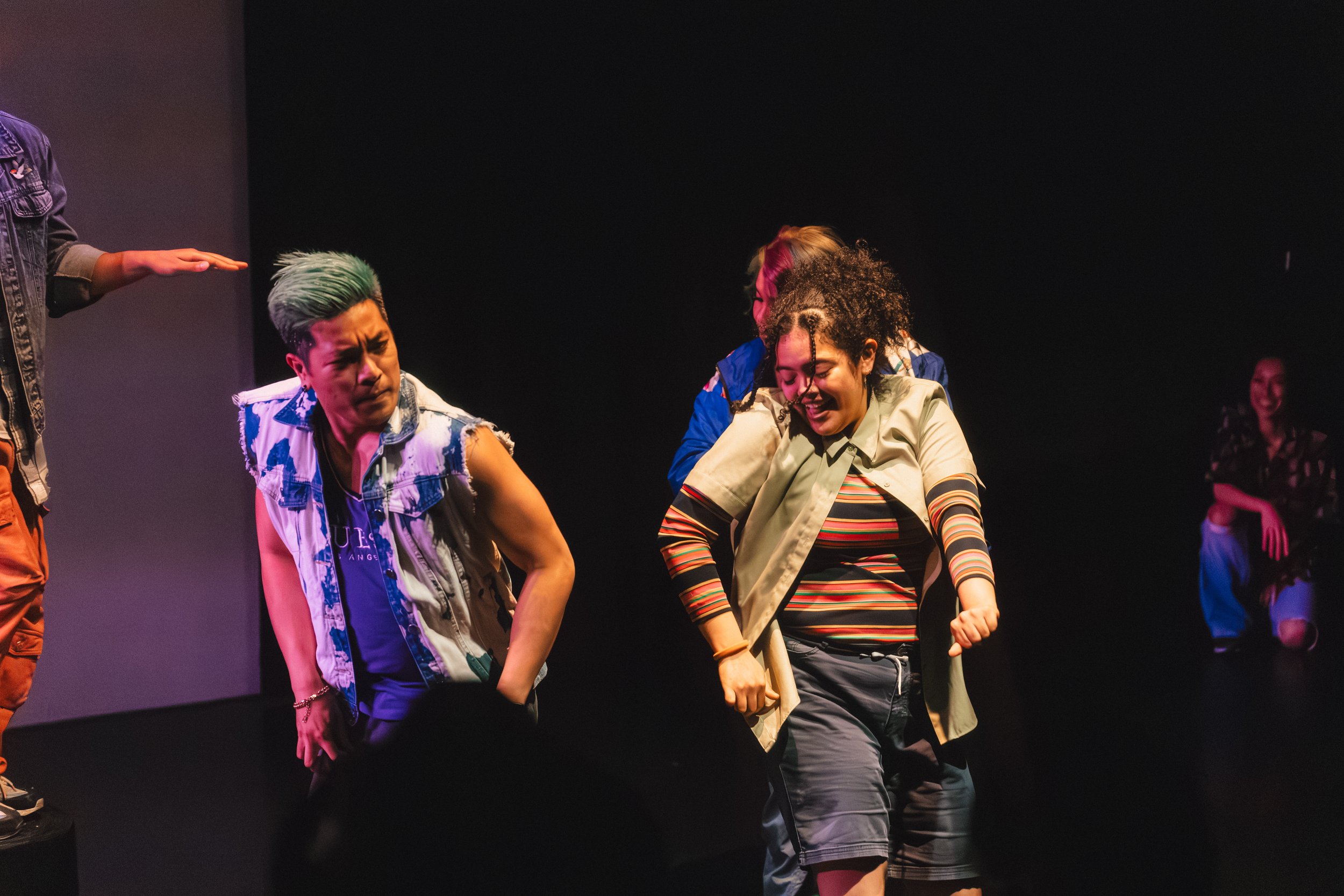 Group of young people dancing and smiling on a stage, with some holding hands.