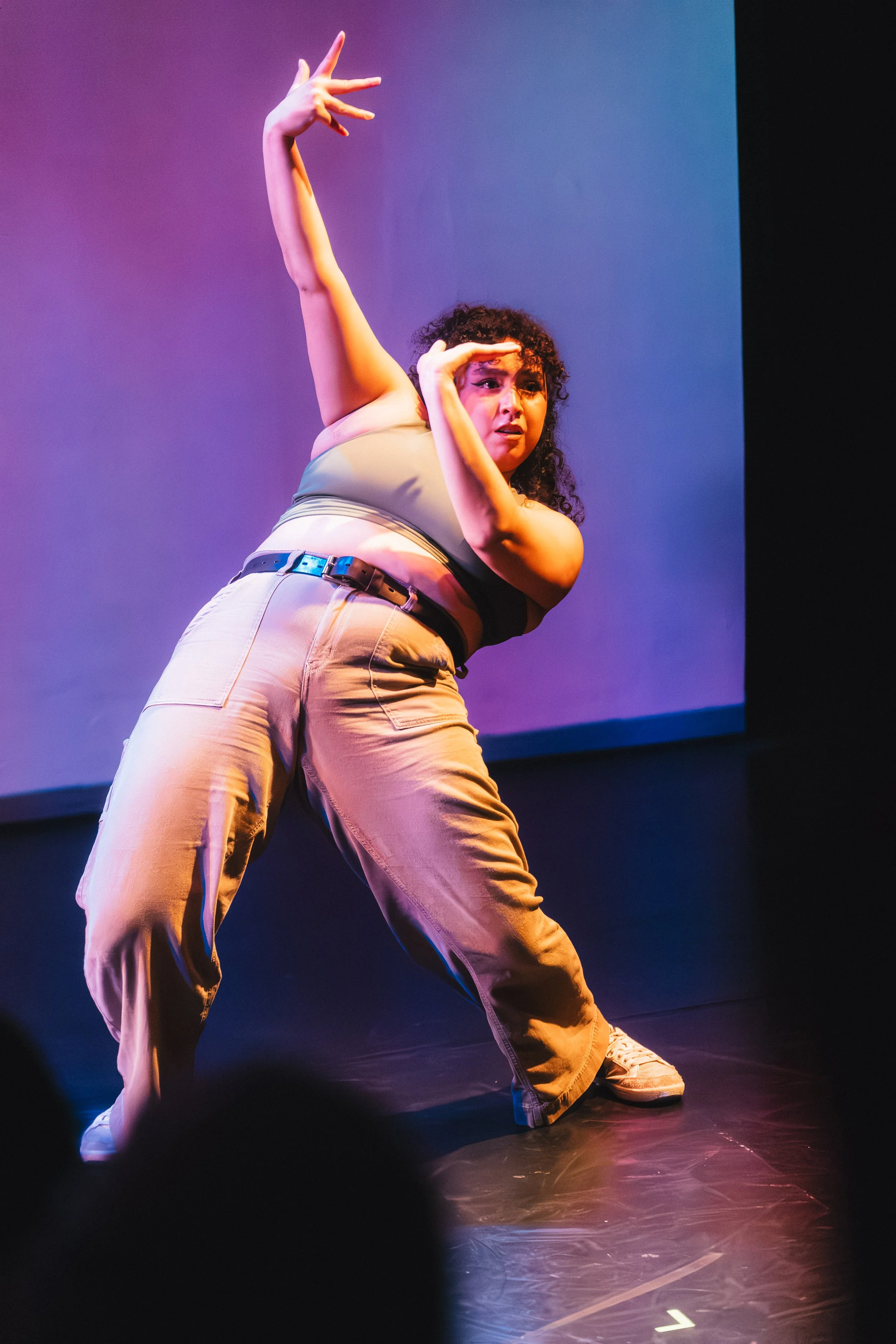 A woman dancing on stage, wearing beige cargo pants, a beige and black tank top, and white sneakers, with colorful stage lighting creating a purple and pink hue.
