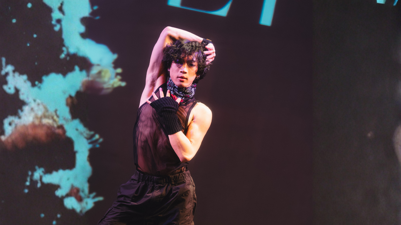 A young man with curly black hair dancing on stage, wearing a black sleeveless shirt, black pants, a black and white bandana, and a black glove on his left hand, with a dark background and colorful abstract graphics.