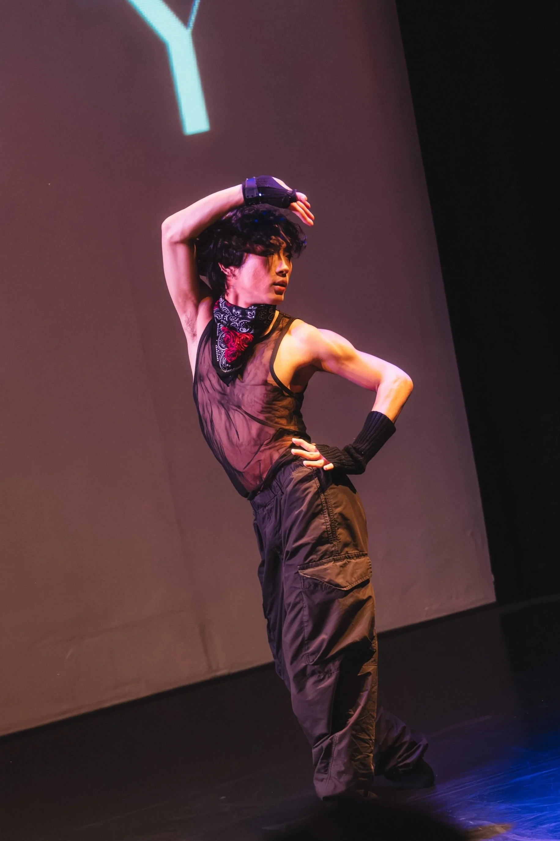 Person with curly hair in black clothing and a bandana around their neck, raising their right arm over their head, standing on a stage with a dark background and a projection of a Y-shaped shape above them.