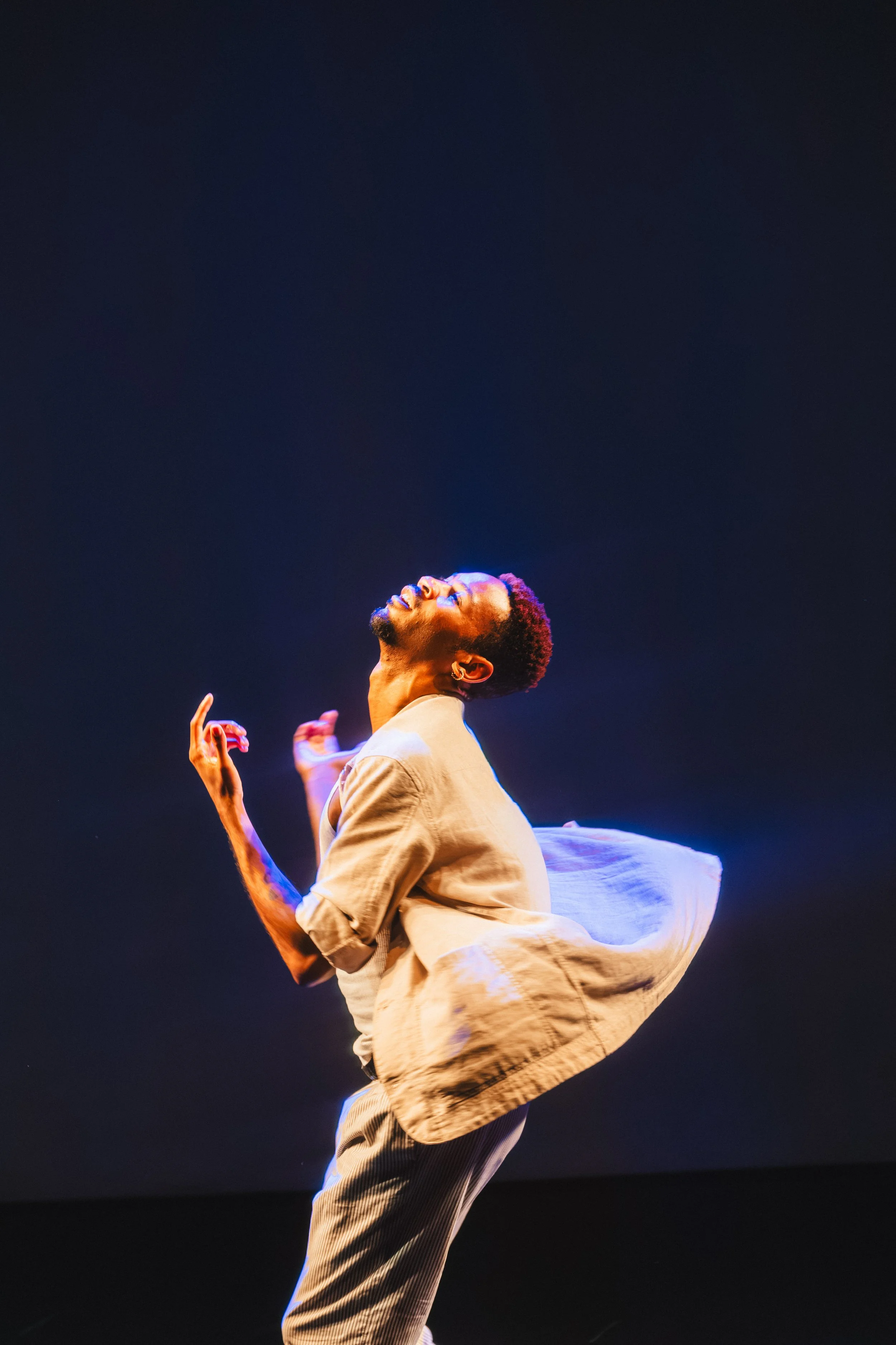 A young man with curly hair and a beard is dancing or performing, looking upward with his eyes closed, wearing a beige jacket and striped pants, against a dark background with stage lighting.