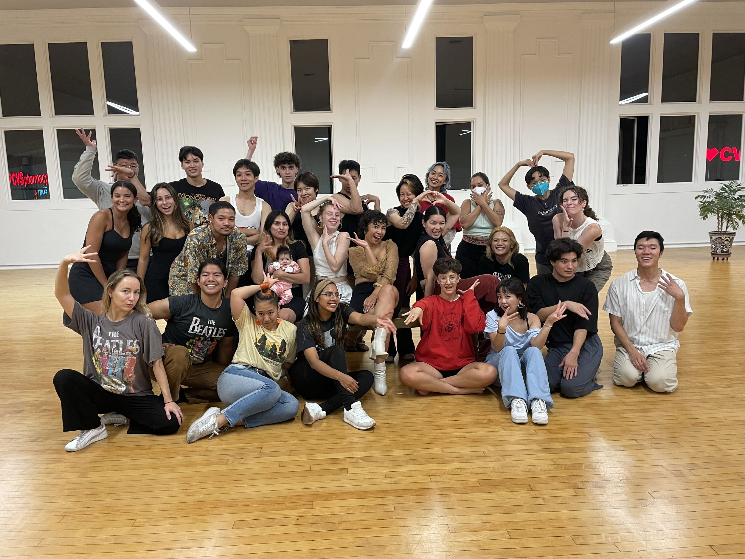 A large group of diverse people are gathered indoors in front of a white wall with multiple large windows, smiling and posing for a group photo. Some are sitting, some are standing, and several are making playful gestures.
