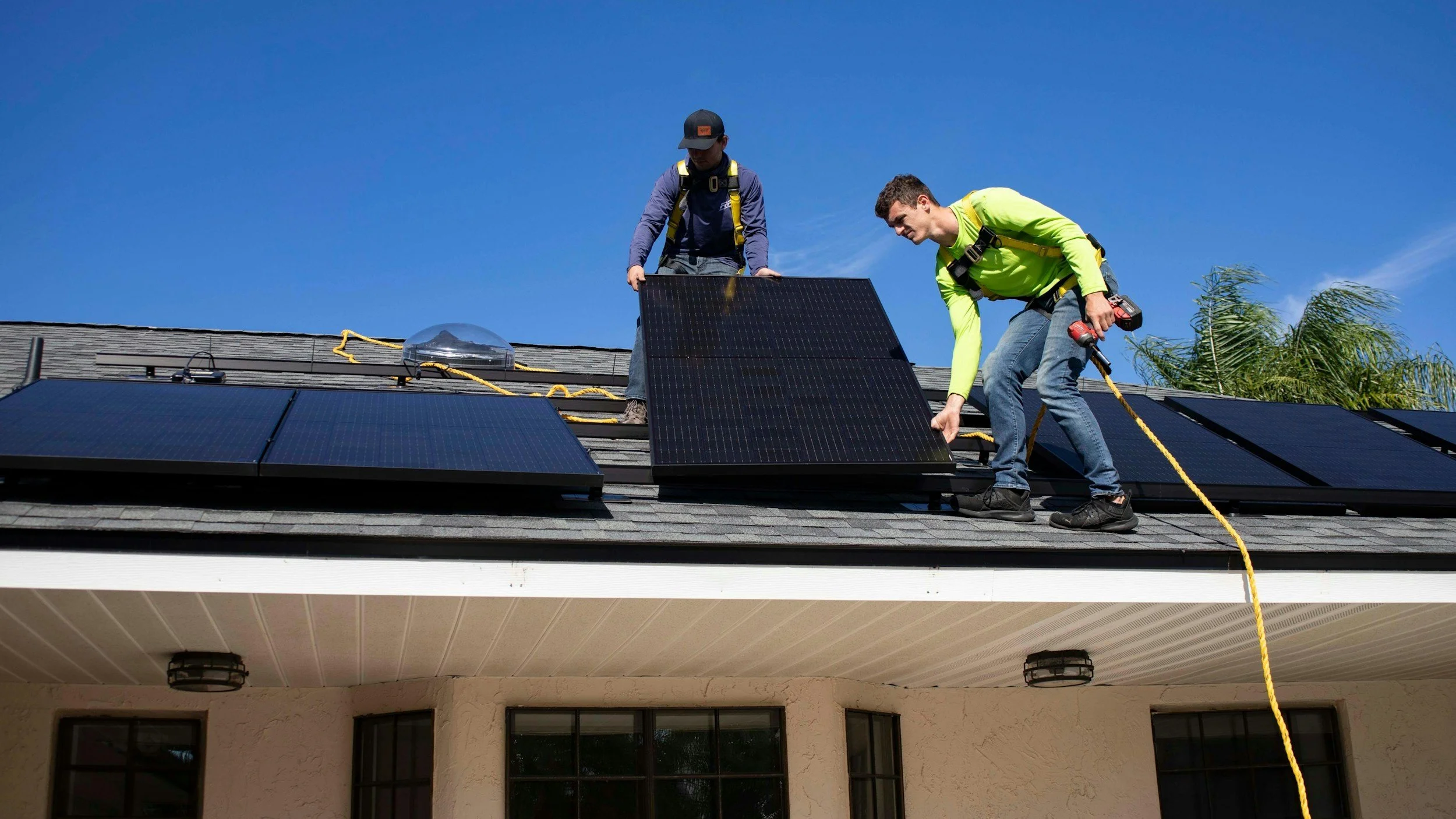 Two workers installing solar panels on a house roof under a clear blue sky.
