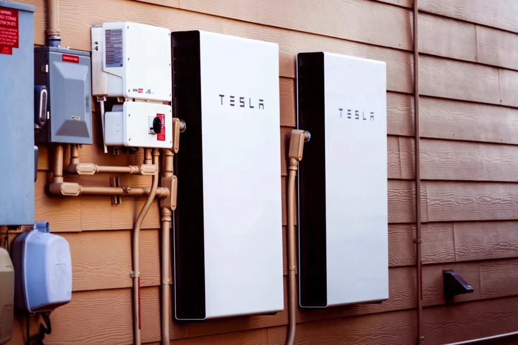 Two Tesla Powerwall energy storage units mounted on a wooden exterior wall, with electrical boxes and conduit pipes nearby.