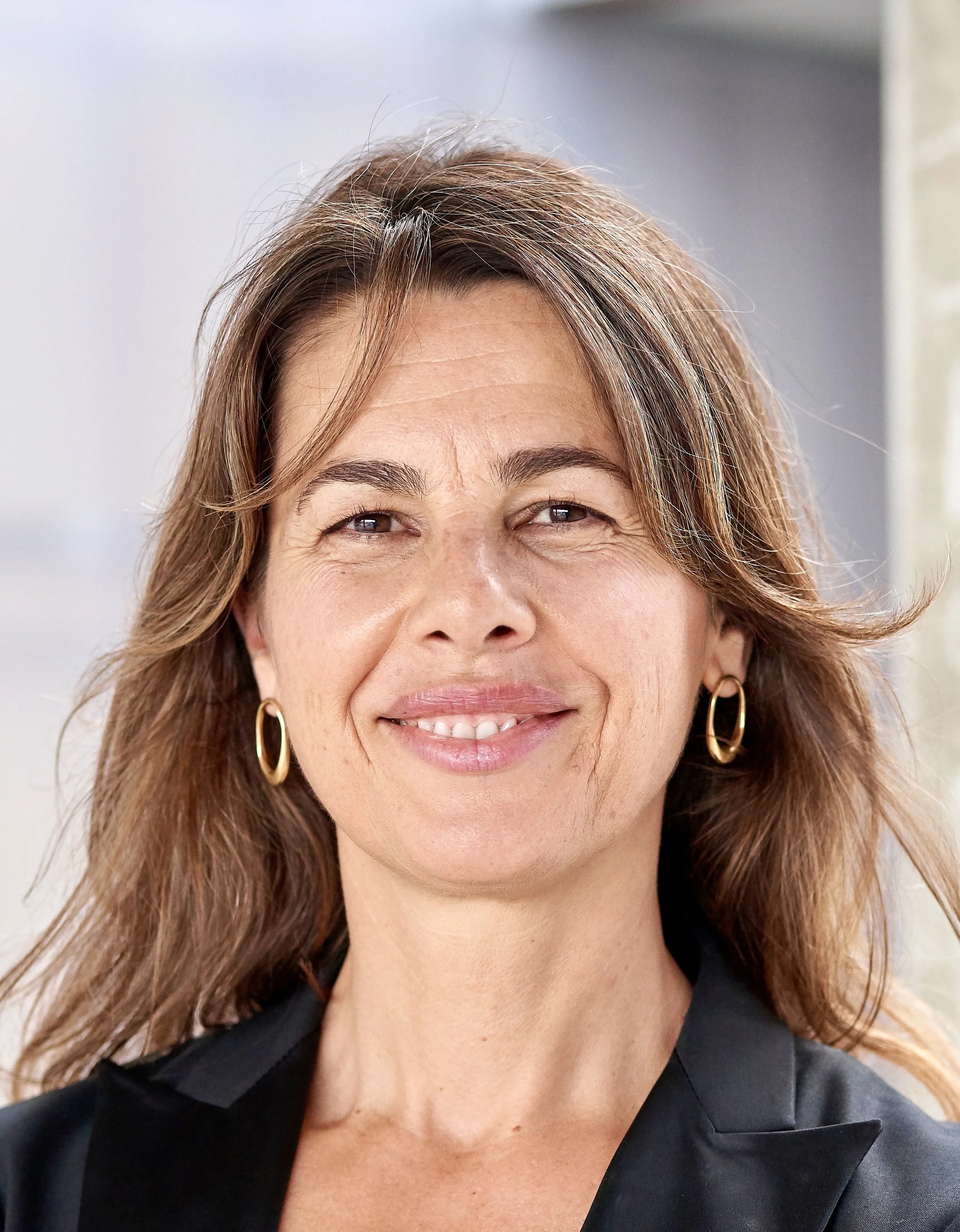 Close-up portrait of a middle-aged woman with shoulder-length brown hair, wearing gold hoop earrings and a black top, smiling in a well-lit indoor setting.