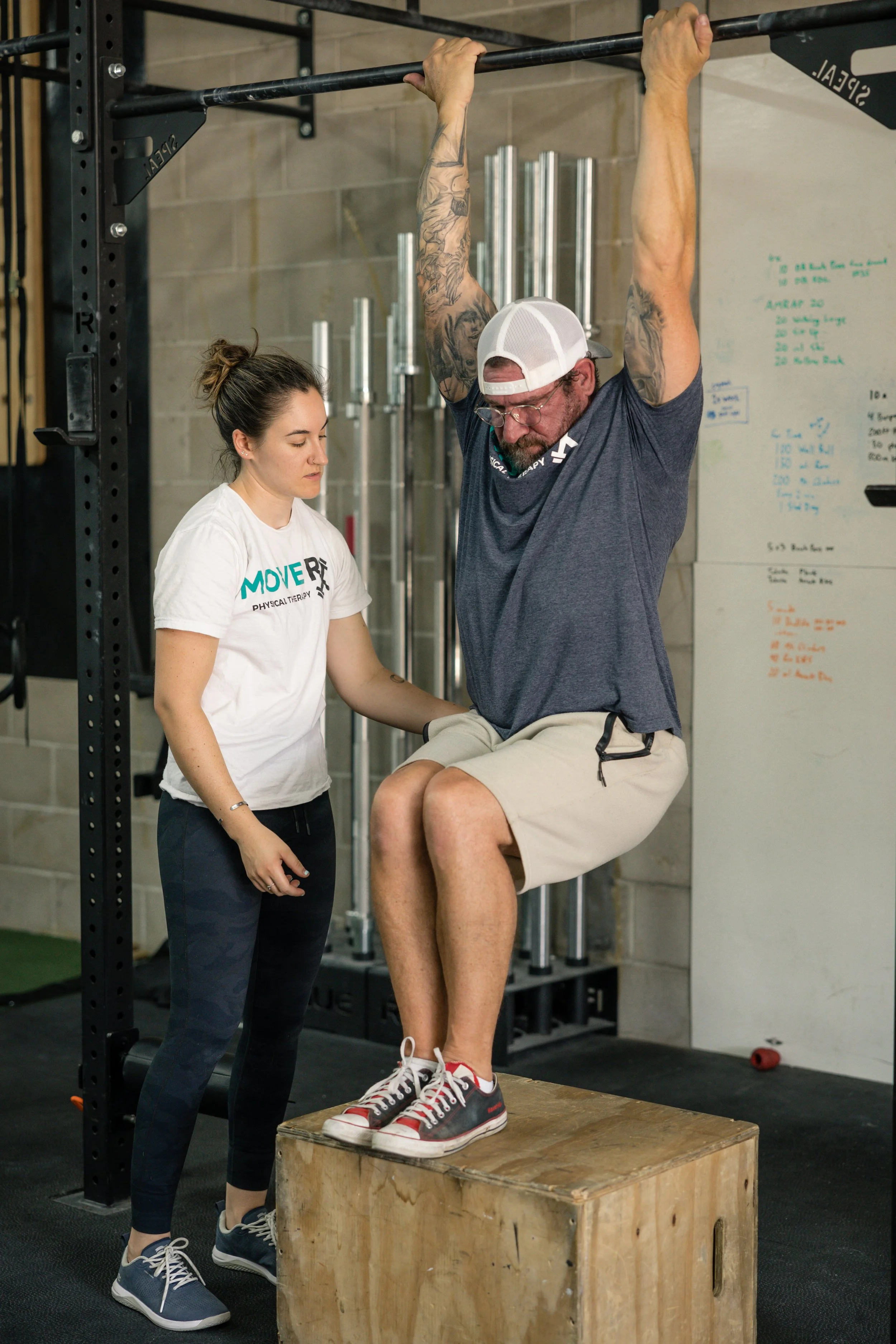 A man performs an upper body stretching exercise on a wooden box while a physical therapist assists him in a gym.