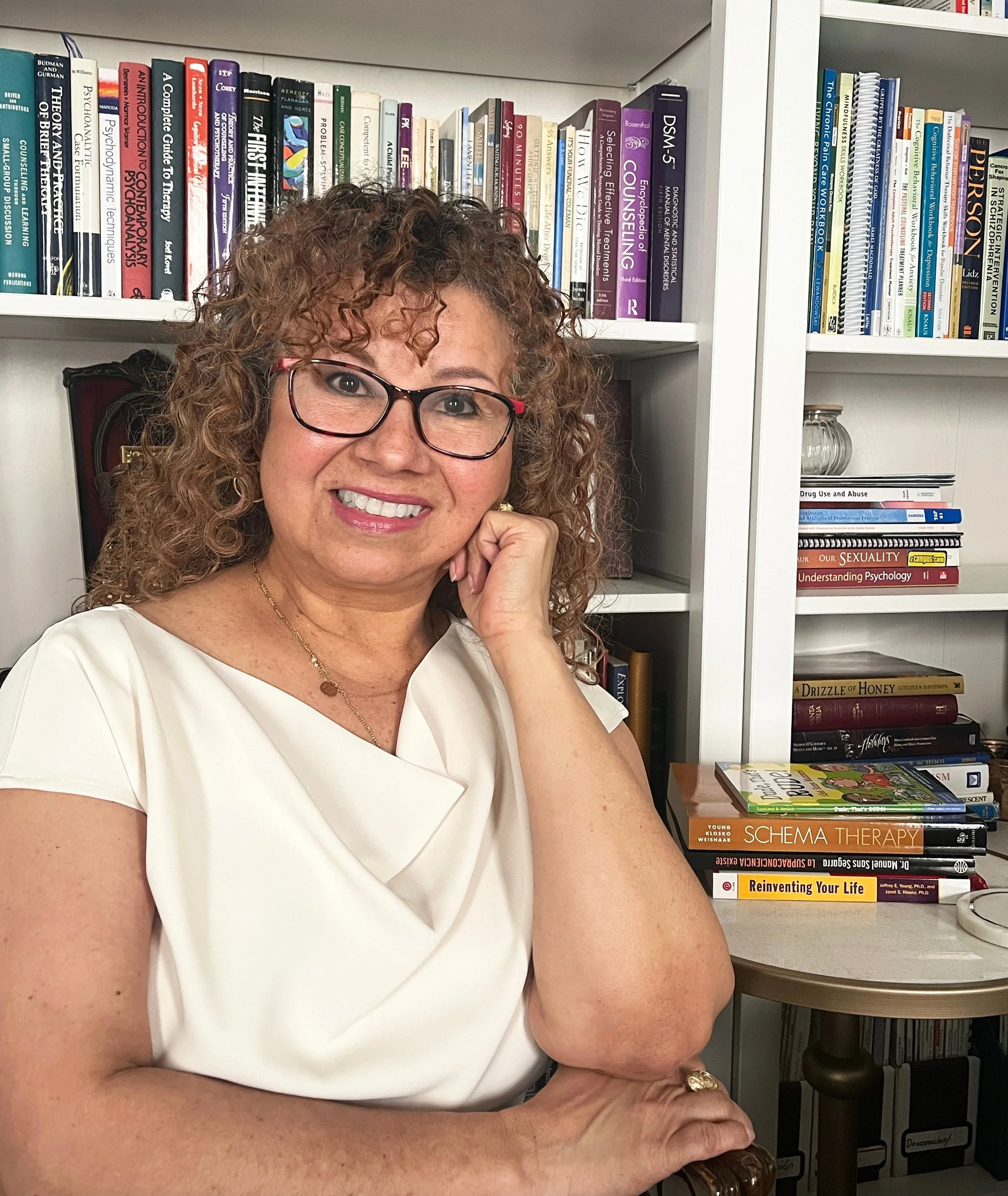 A woman with curly hair, glasses, and a white top, smiling and resting her chin on her hand, sitting in front of a bookshelf filled with various books on psychology, therapy, and self-help, on a table.
