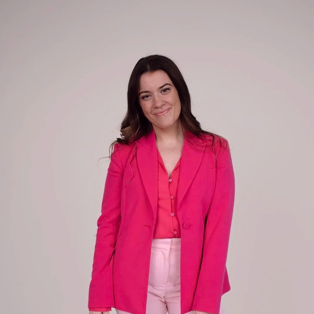 A woman with long dark hair wearing a pink blazer, pink shirt, and light pink pants standing against a plain white background.