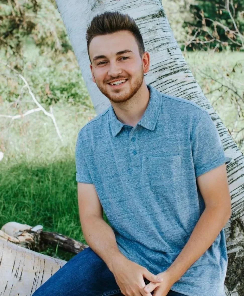 A young man with short brown hair and a beard, smiling while sitting outdoors next to a tree with white bark, in a natural setting with green grass and trees in the background.