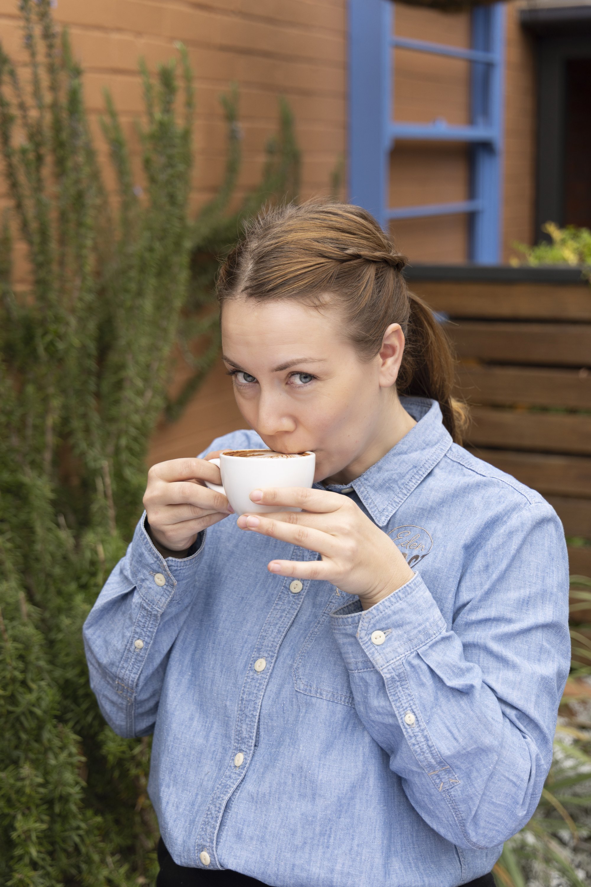 A woman with brown hair in a braid, wearing a blue denim shirt, drinks from a white cup outdoors in a garden setting.