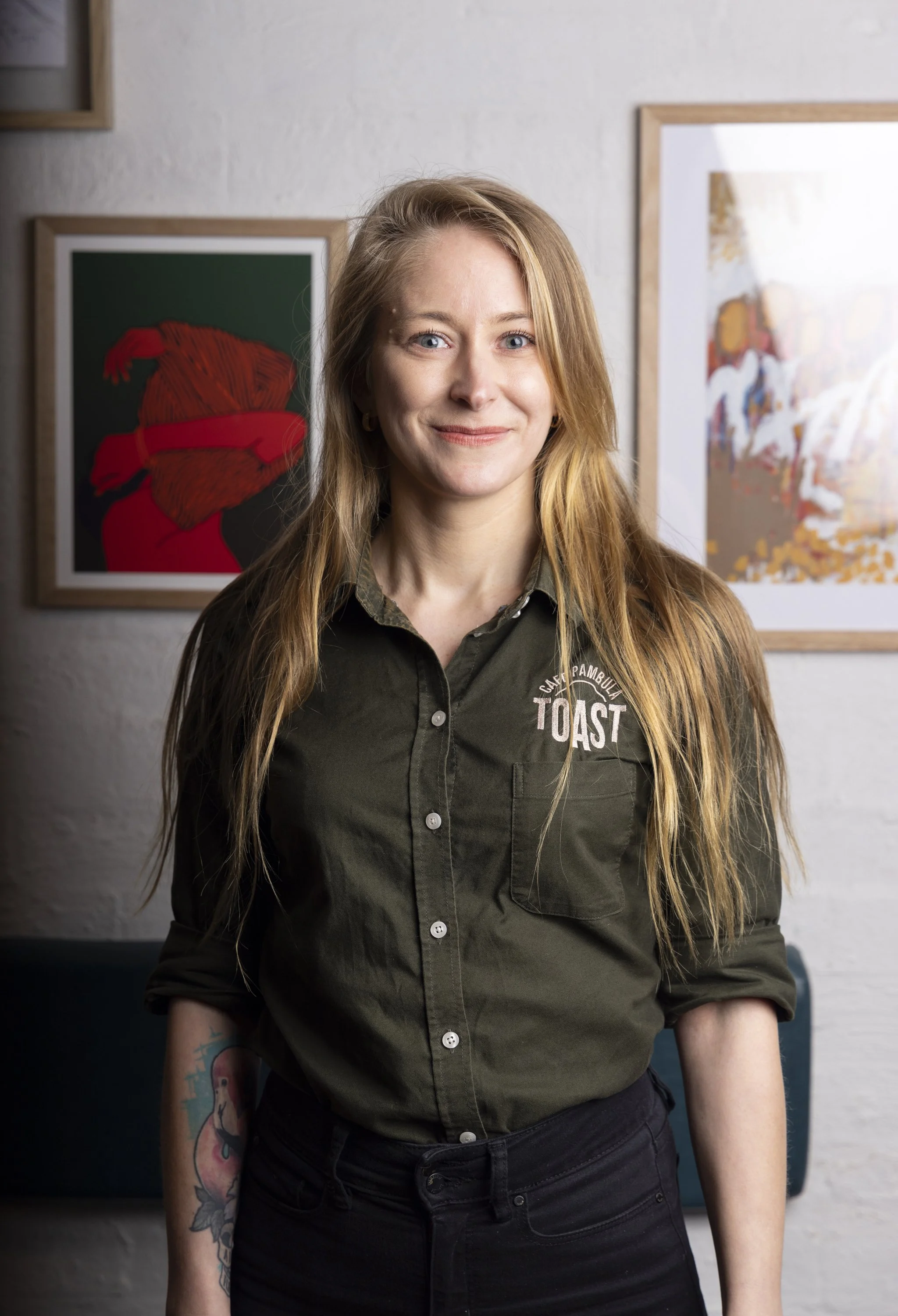 A woman with long red hair and a tattoo on her arm, wearing a dark green shirt with "TOAST" embroidered on it, standing in front of framed artwork on a white wall.