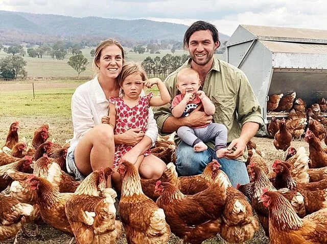 Family of four sitting with chickens on a farm, smiling at the camera.