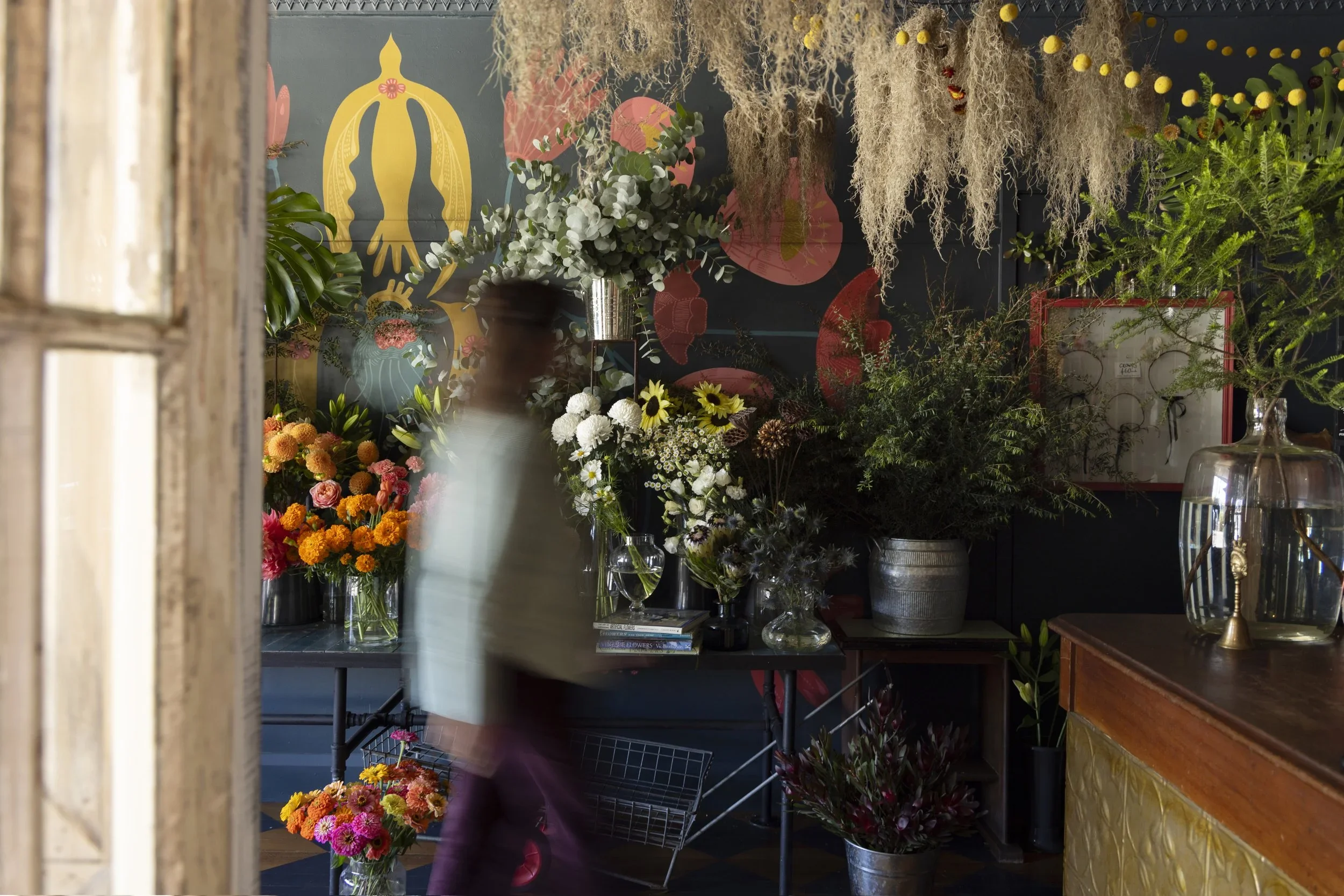 Interior of a flower shop with various colorful flowers and greenery on display, a blurred person walking past, and artworks on the dark wall in the background.
