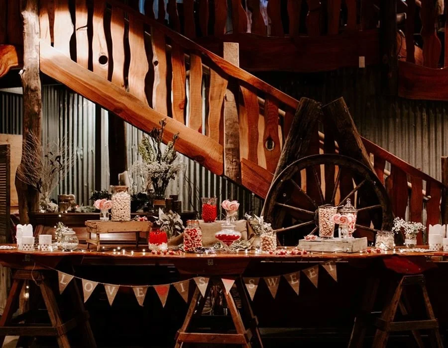 A decorated table with jars of candies and sweets, surrounded by floral arrangements and candy-themed decorations, set against a rustic wooden staircase and wagon wheel in the background.