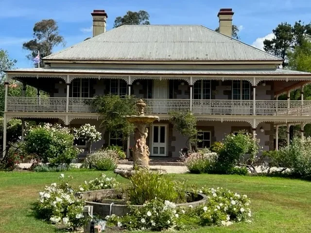 Historic house with a large wraparound porch, chimney stacks, surrounded by a garden with a fountain and rose bushes, on a sunny day.