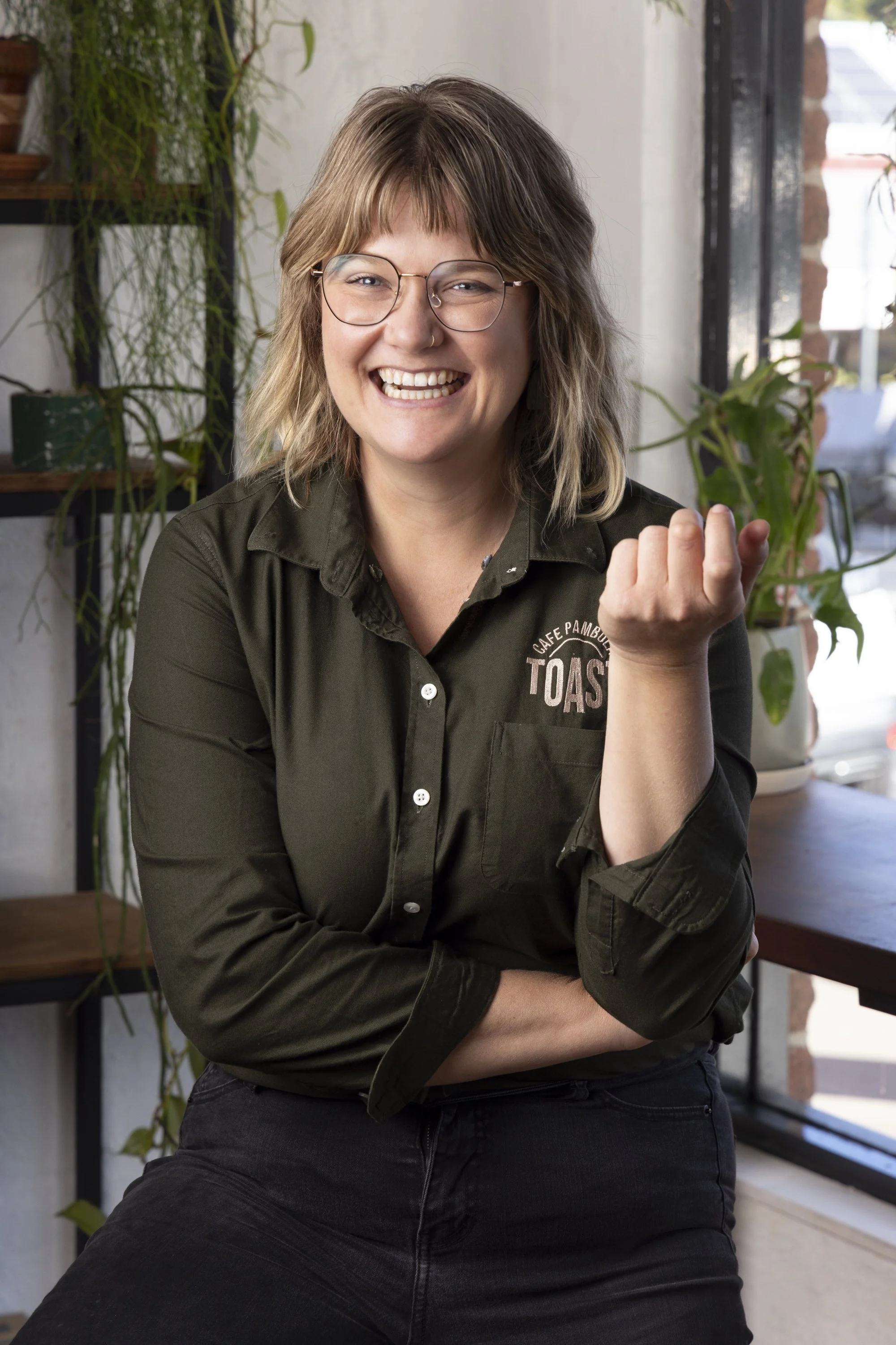 Smiling woman with glasses and a nose ring, wearing a black shirt with a restaurant logo, sitting indoors near a window with plants in the background.