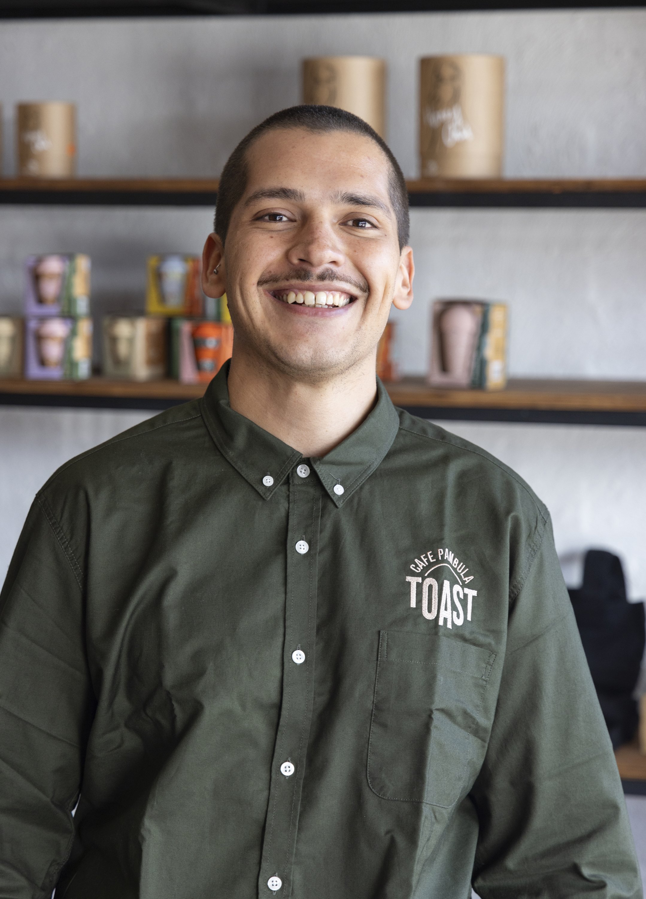 A smiling man in a dark green button-up shirt standing in a coffee shop. The shirt has a logo that reads 'Cafe Panbula Toast.' Shelves with colorful coffee cups are visible in the background.