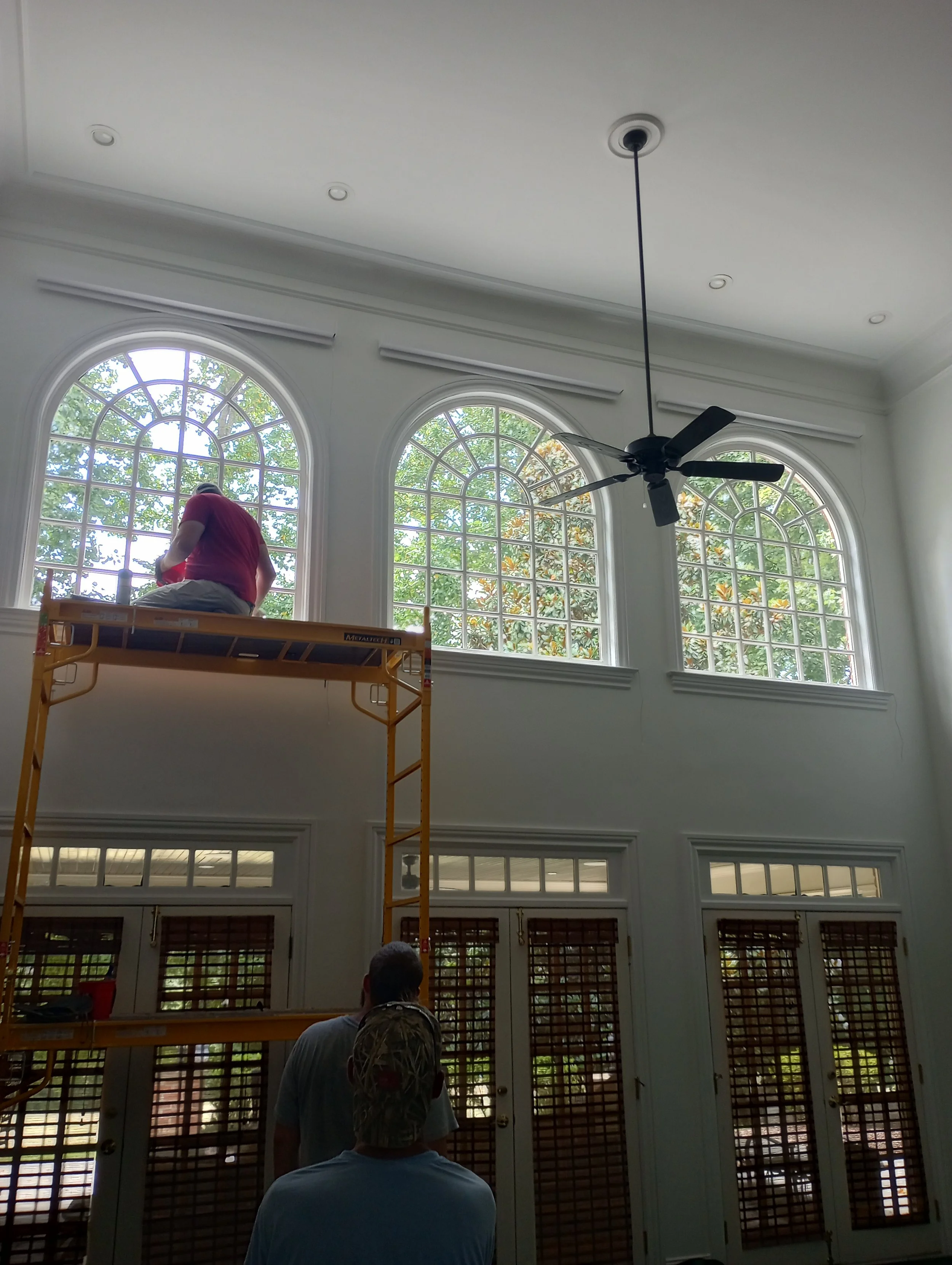 Two workers are installing or cleaning brown window shutters inside a room with large arched windows and a black ceiling fan. One worker is on a yellow scaffolding, and the other is standing below, looking up.