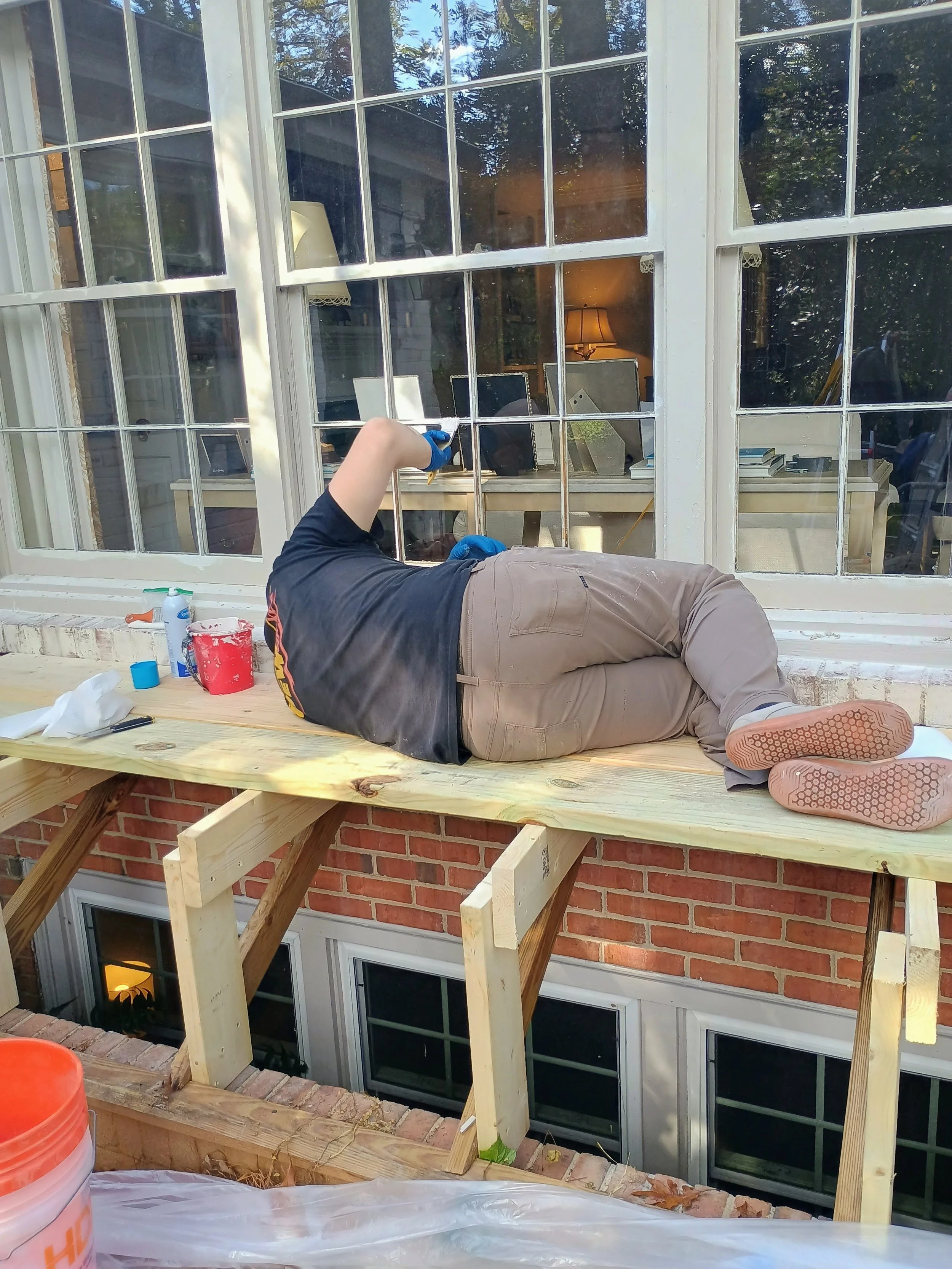 Person lying on a wooden platform under construction in front of a large window, working on the window frame with tools and supplies nearby.