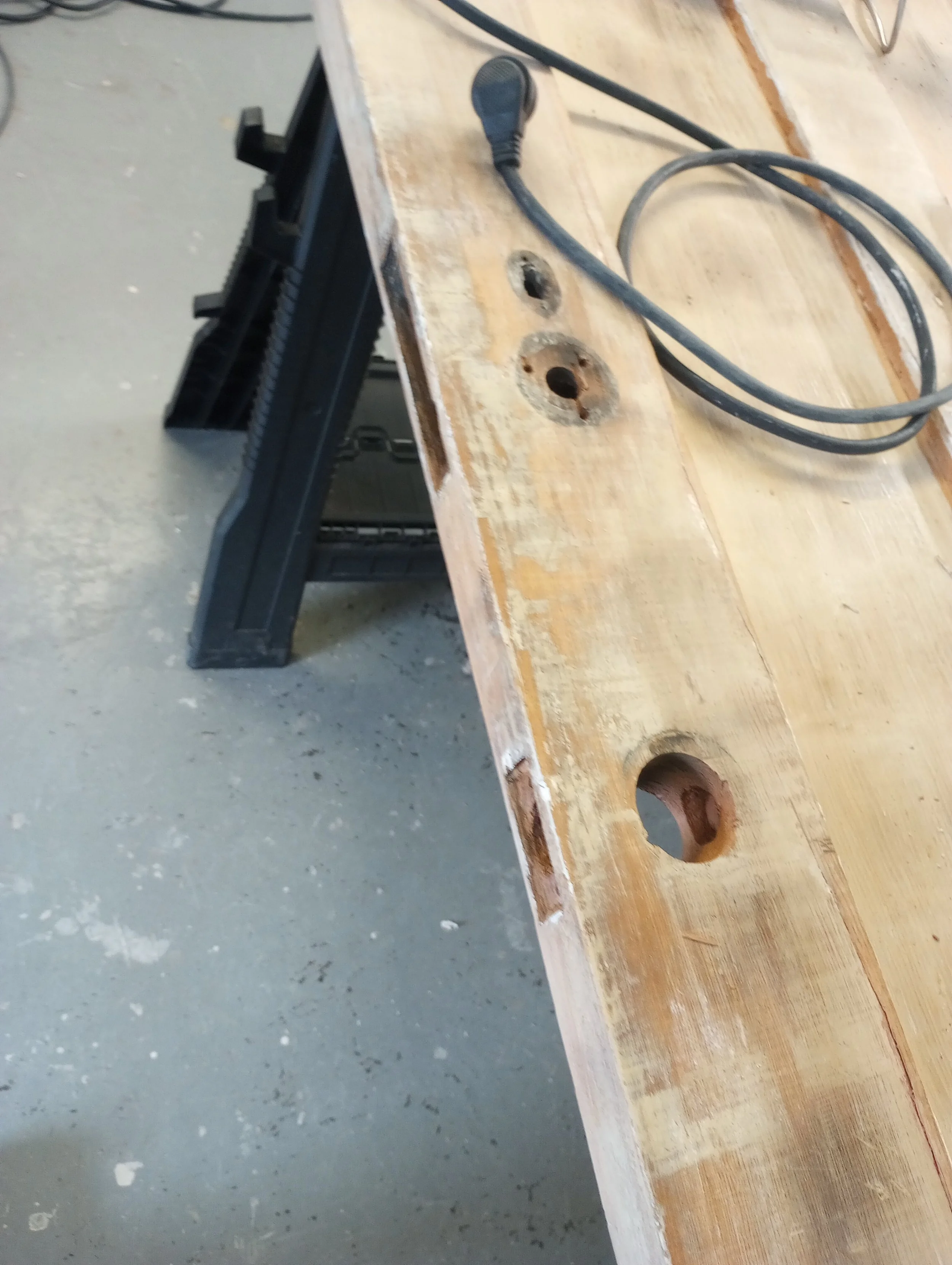 Close-up of a wooden workbench with three drilled holes and a black clamp on the side, with electrical cords on the surface.
