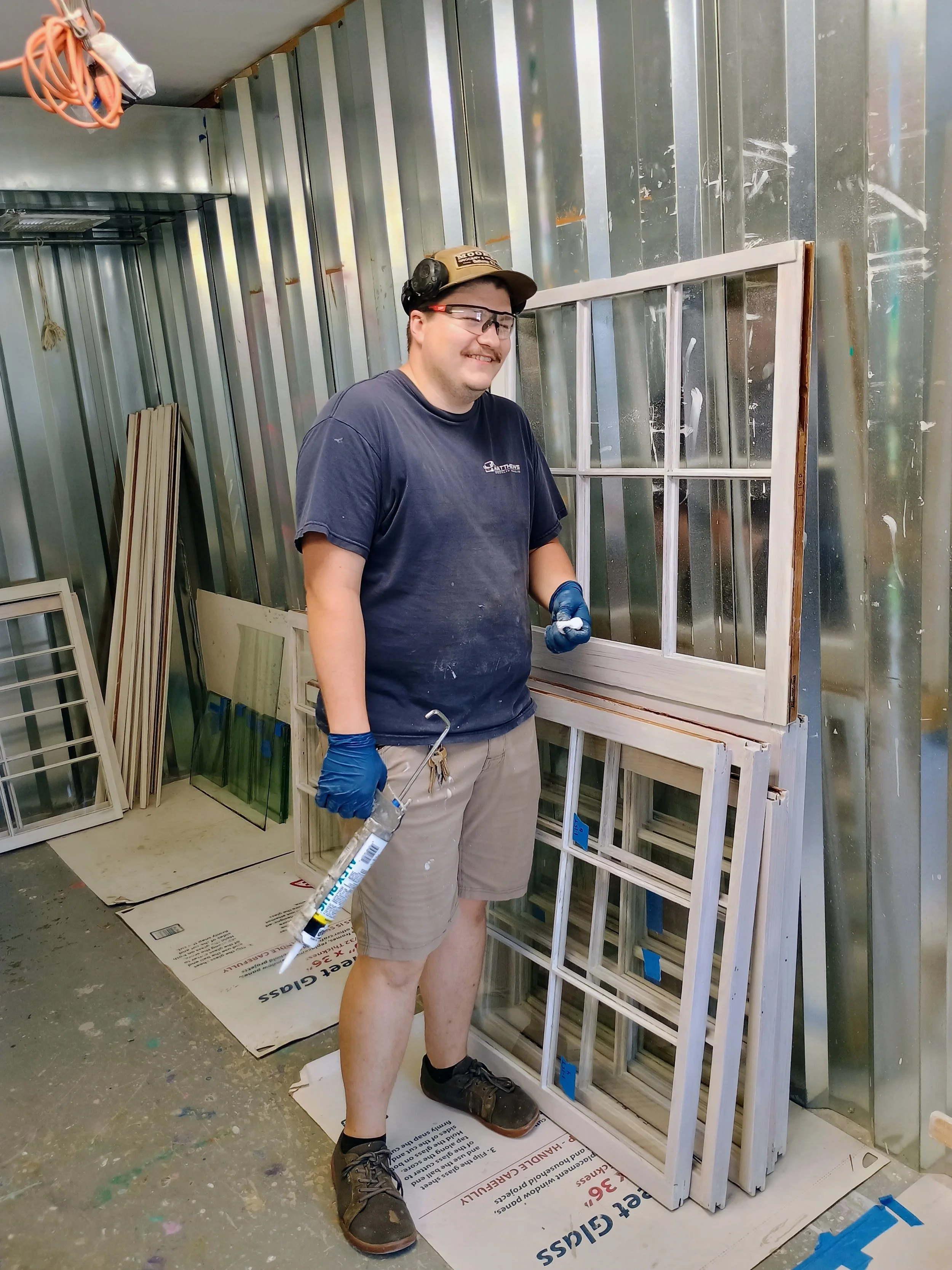 A man wearing a dark blue t-shirt, beige shorts, black work shoes, safety glasses, and a baseball cap is standing in a workshop holding a caulking gun in his right hand. He is smiling and standing next to a wooden window frame, with other window fram