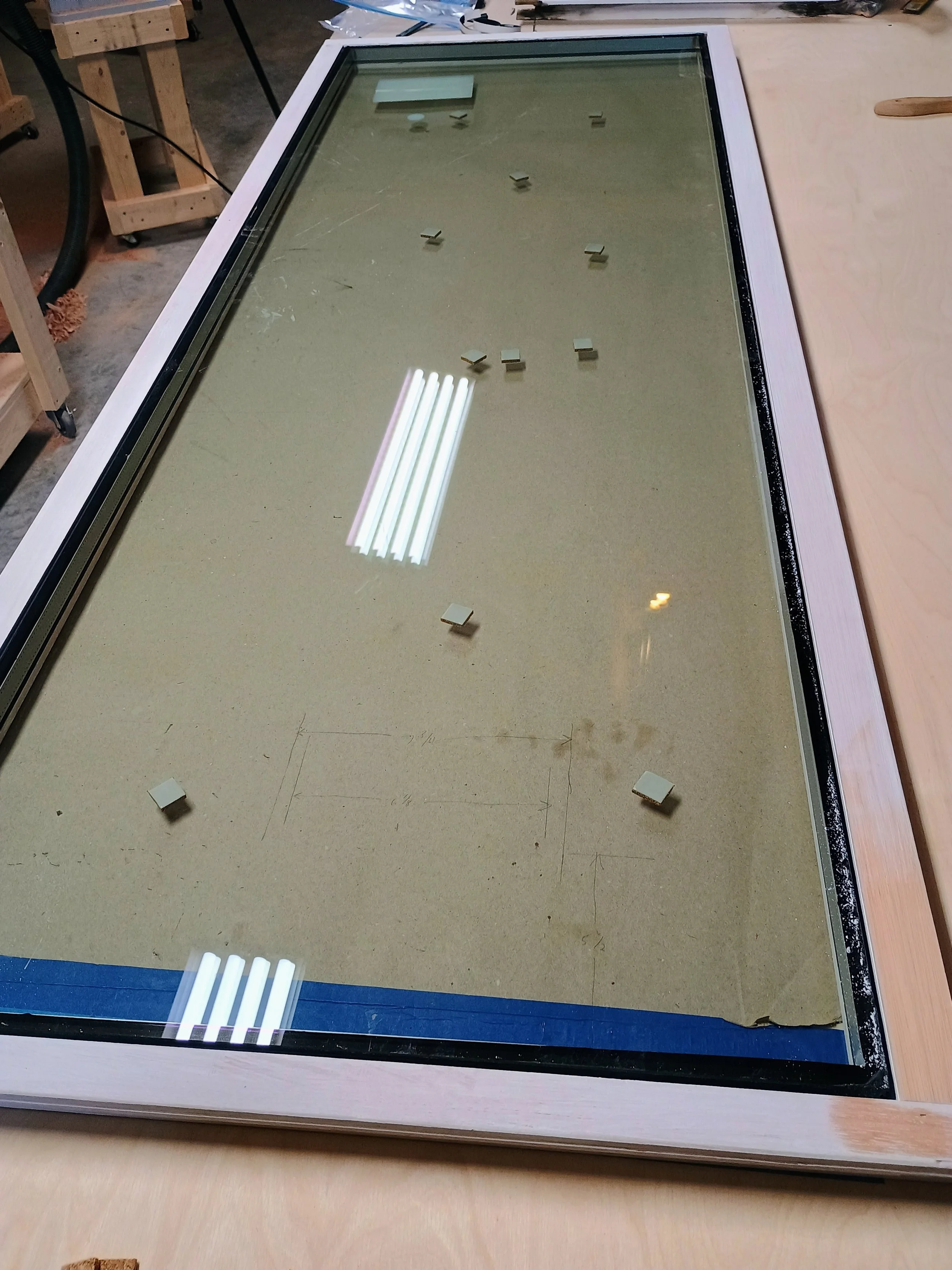 A rectangular window with a white frame and glass pane resting on a worktable in a workshop, with several small white blocks placed on the glass surface.