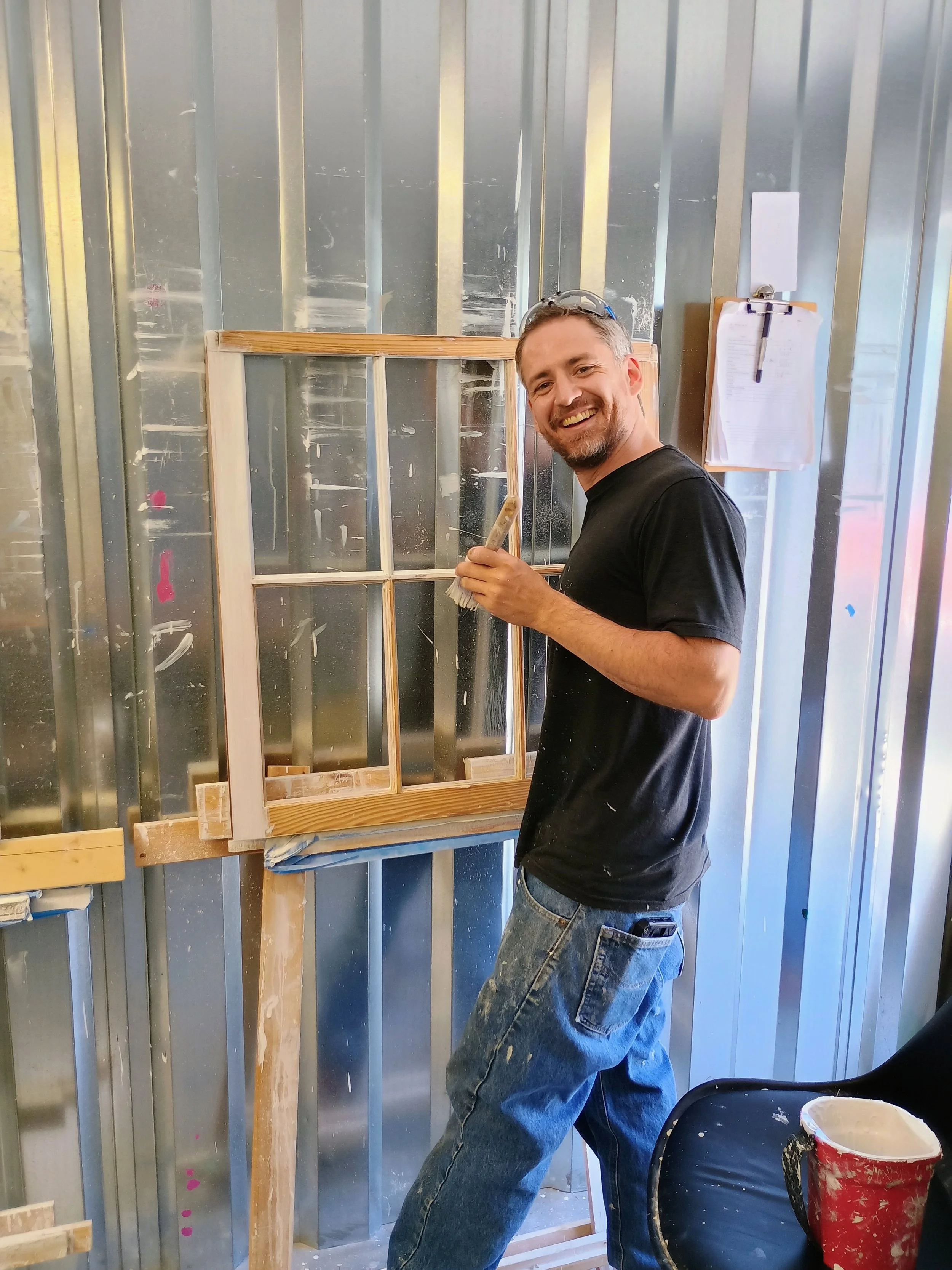 A man working on building a wooden window frame in a workshop, smiling at the camera.