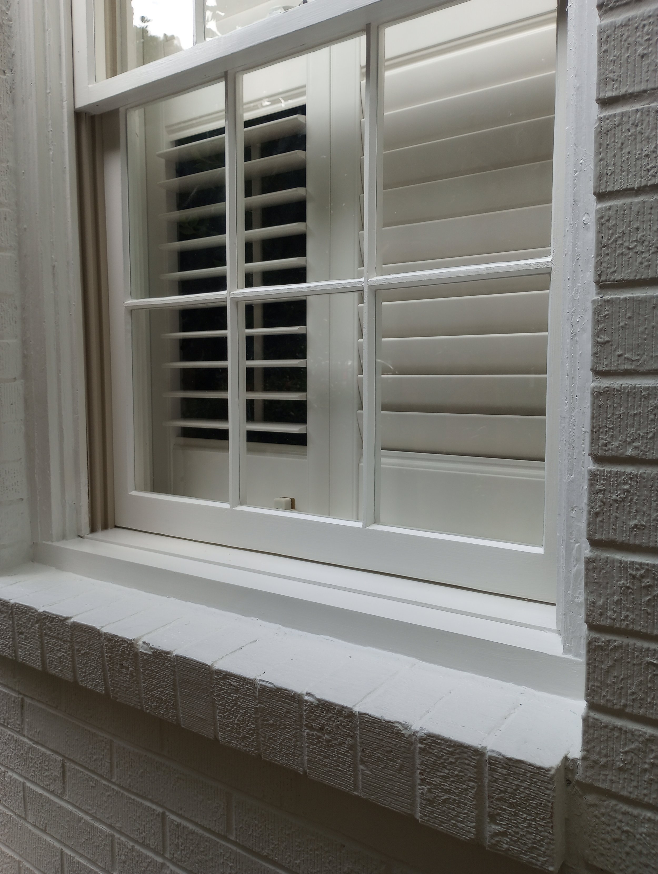 Close-up of a white-framed window with brick exterior wall, white shutters, and decorative brick sill in a house.