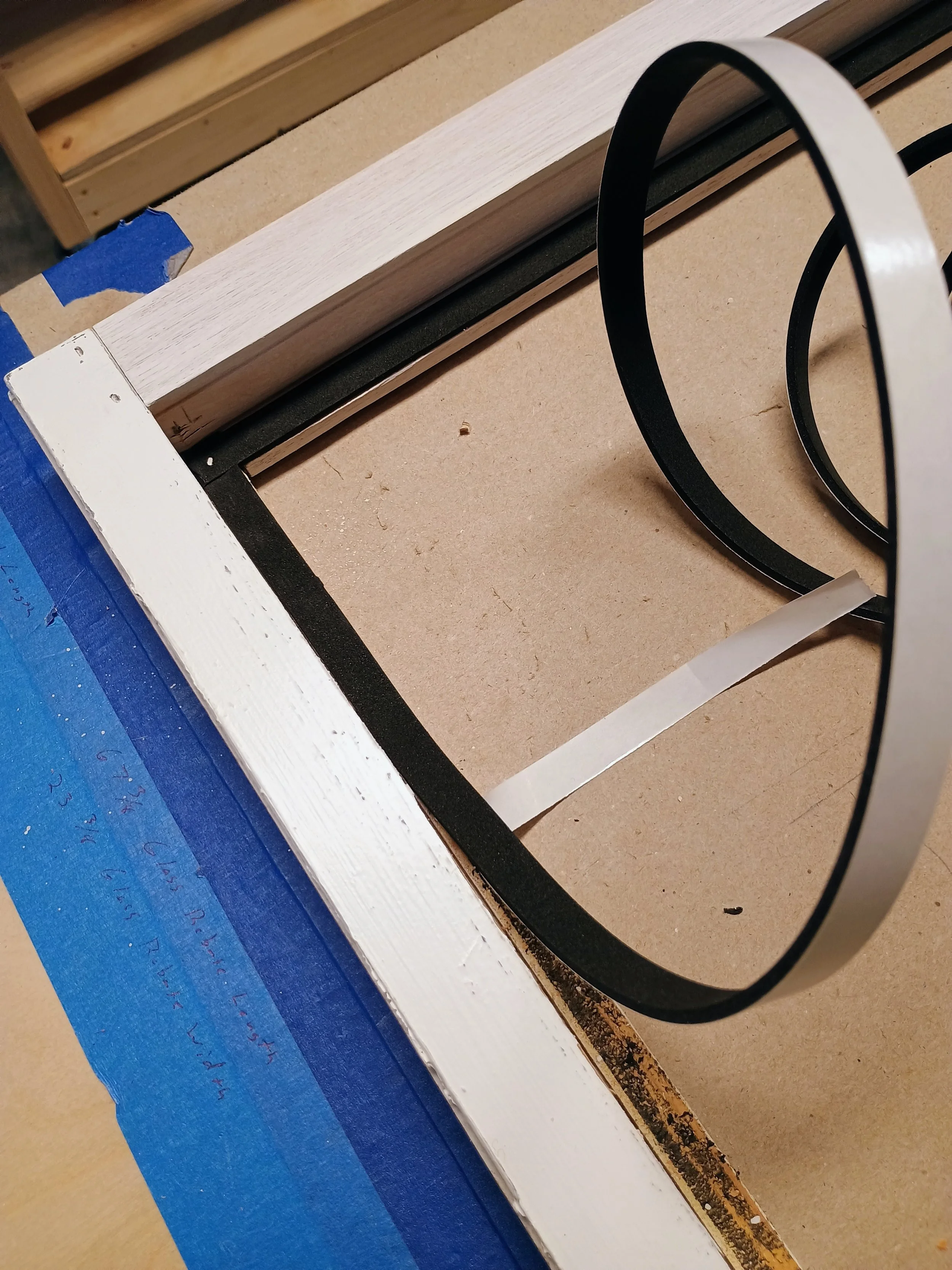 Close-up of a woodworking project with a white painted wooden frame, black metal trim, a piece of blue painter's tape, and a strip of white tape on a brown work surface.
