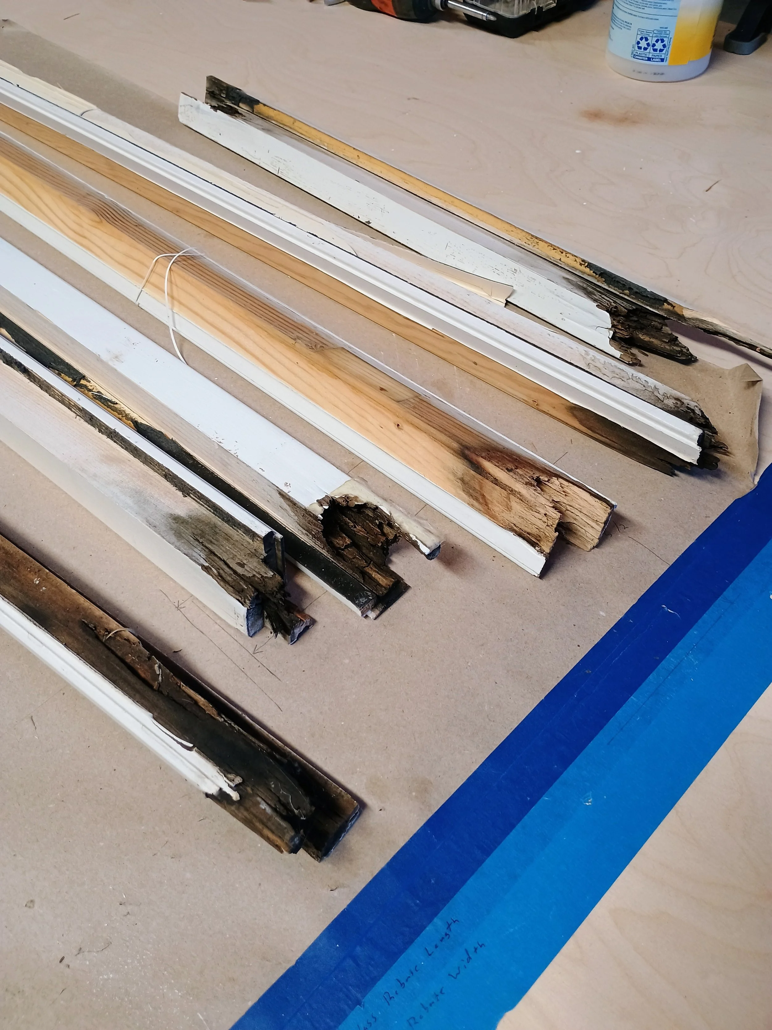 Various pieces of wood and moldings, some burned or damaged, laid out on a workspace with a blue painter's tape marked section.