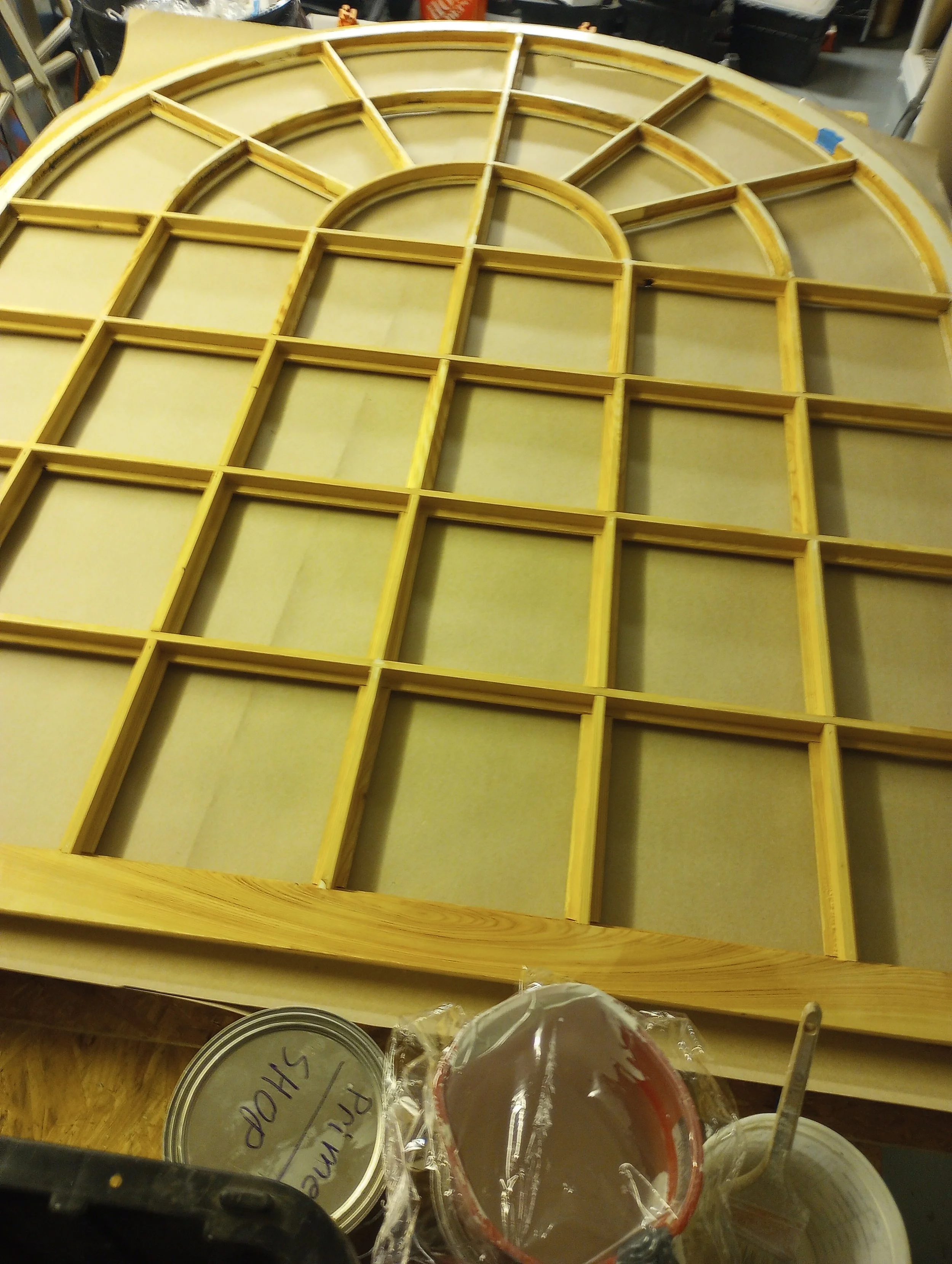 Wooden framework for a window with multiple panes and an arched top, set atop a work table with painting supplies nearby.