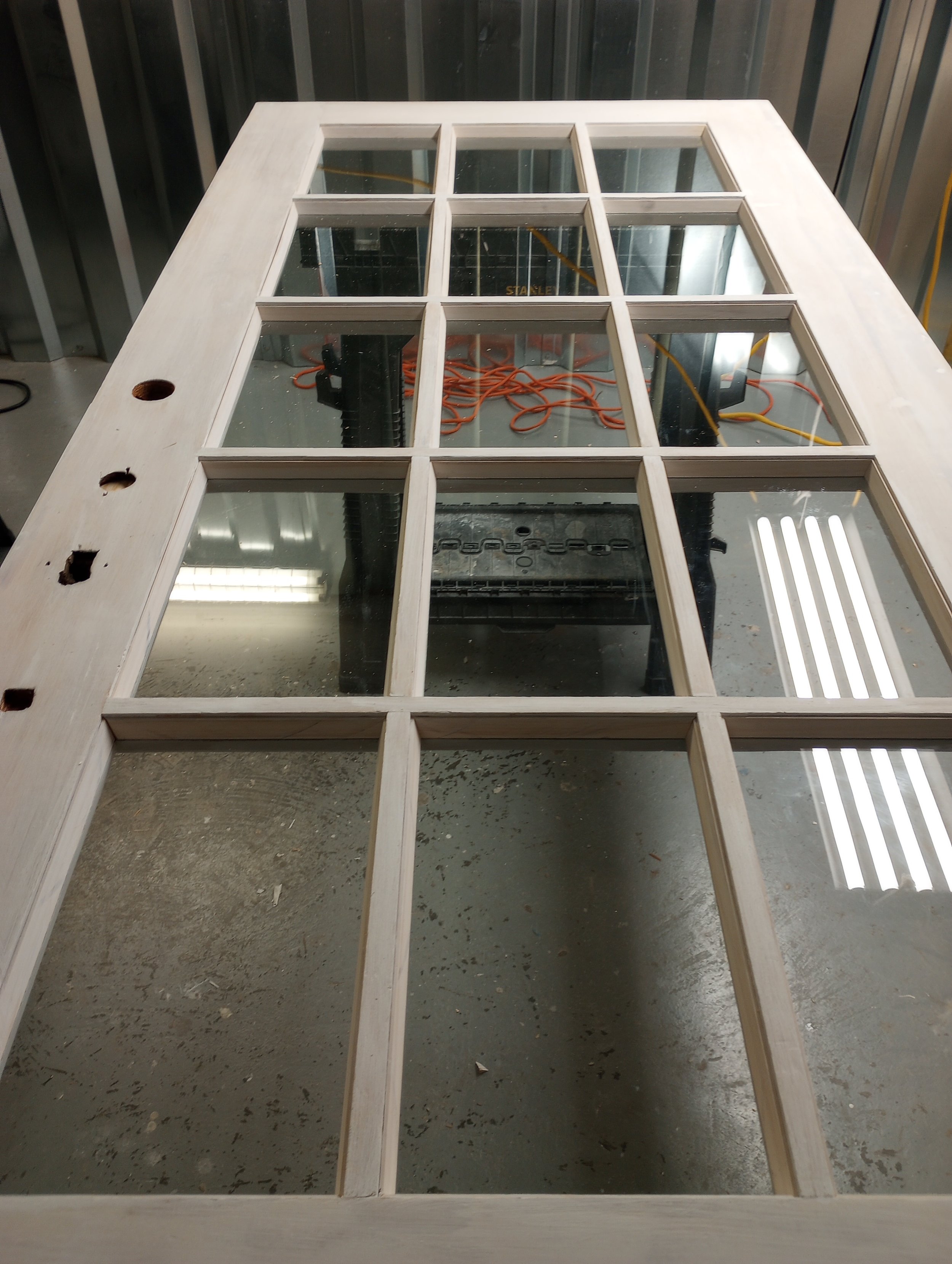 A wooden frame of a door with glass panes laid on a worktable in a workshop, with tools and cords visible underneath.