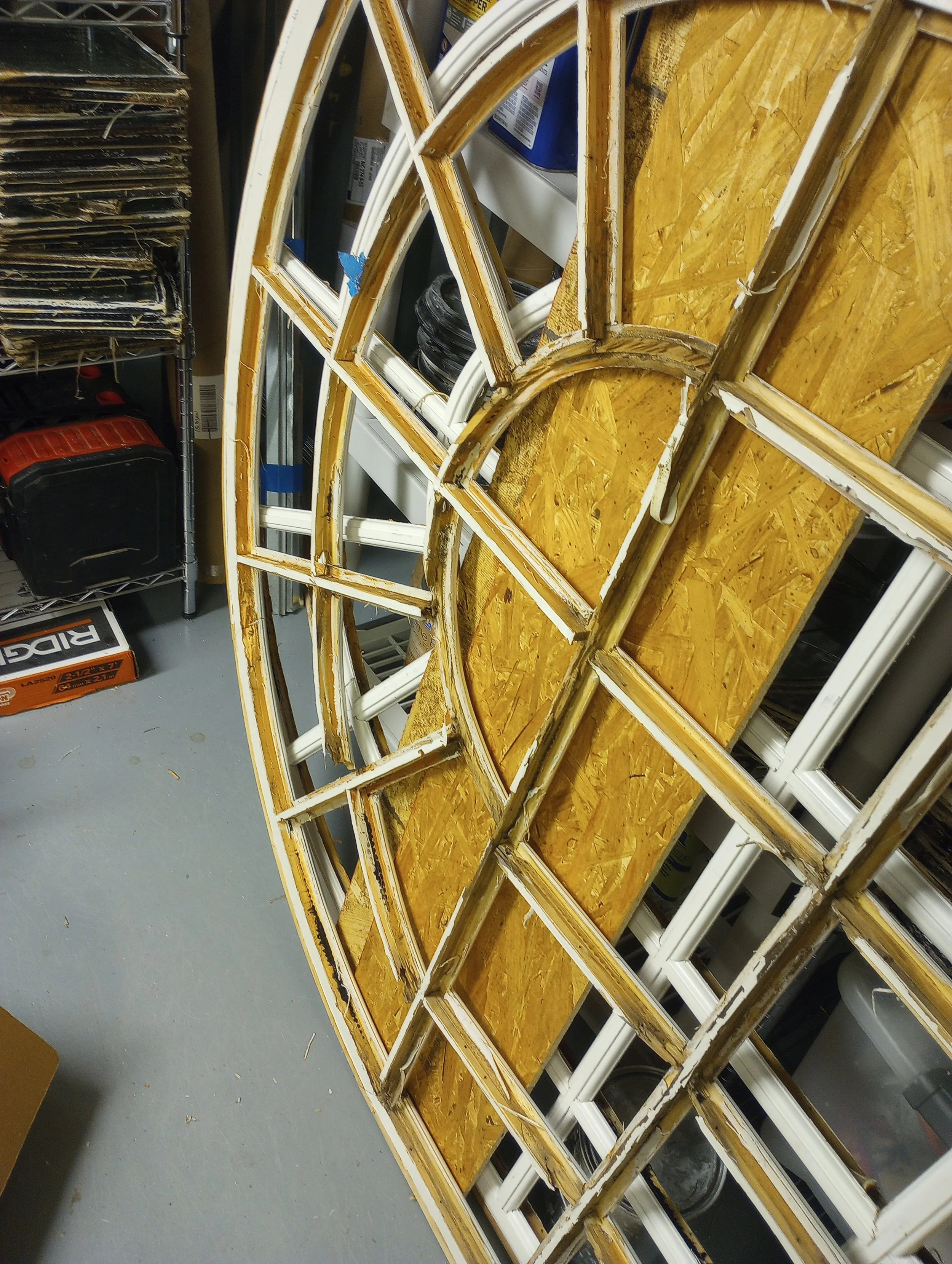 The image shows a partially assembled white metal and wooden shelf or display unit leaning against a storage area with various boxes, containers, and tools nearby.