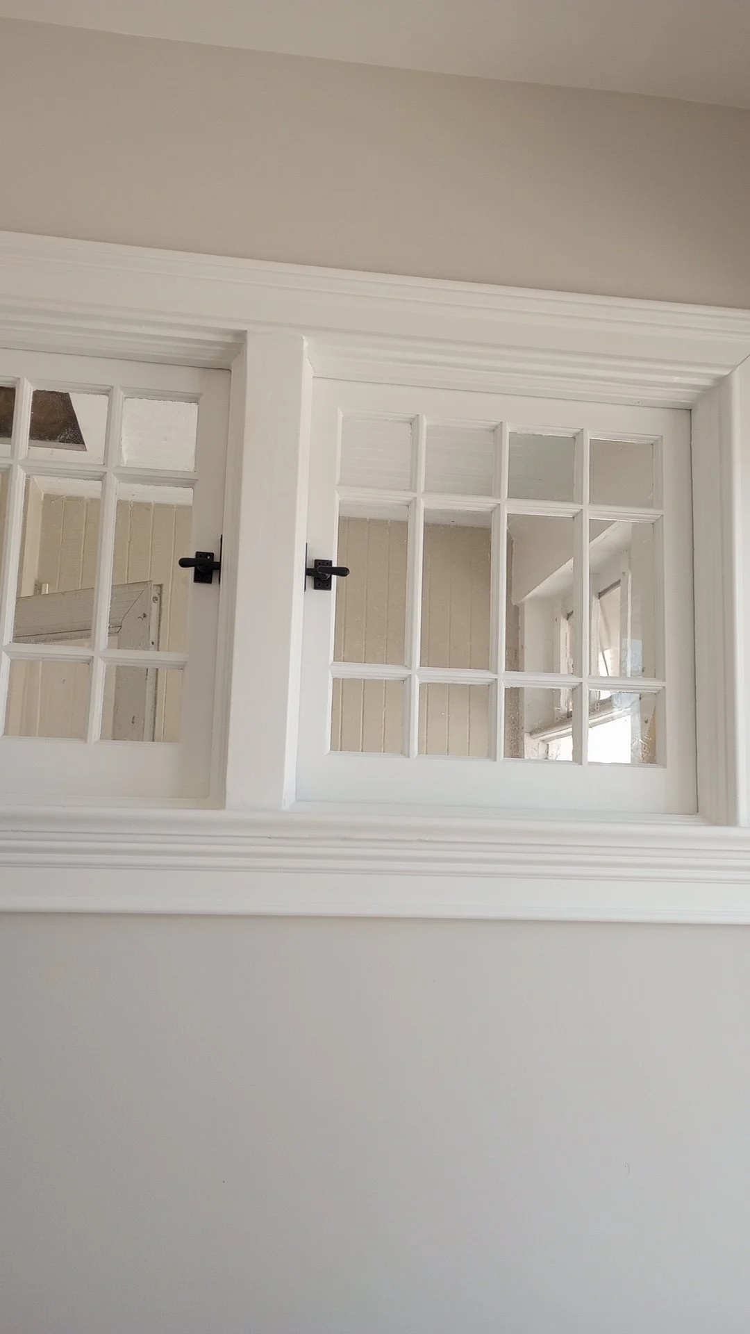 Image of two white framed windows with small glass panes, set in a white wall with decorative crown molding. The windows are slightly open with black latches.
