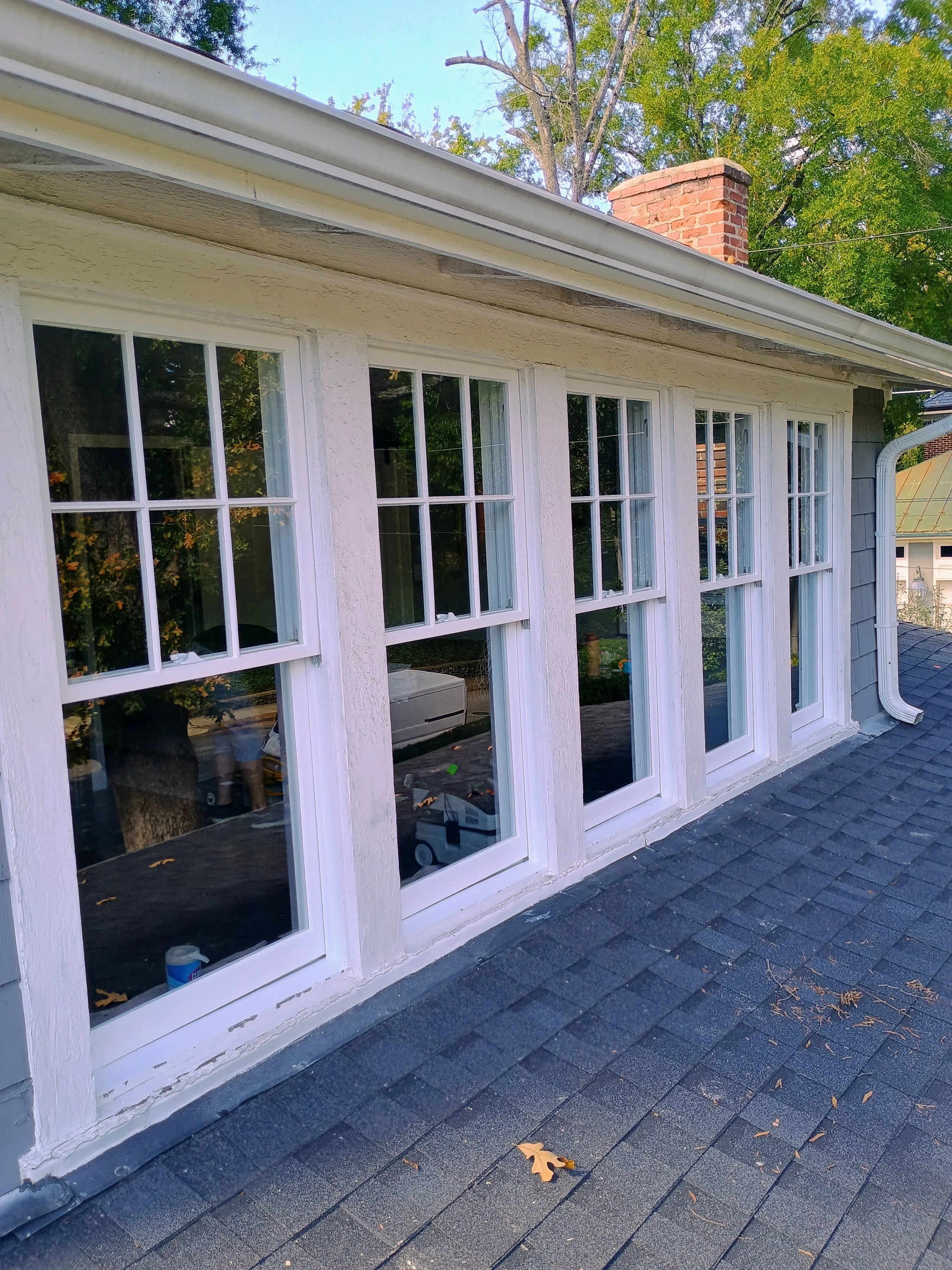 Exterior of a house with five large double-pane windows, white trim, shingled roof, and a brick chimney. Fall leaves are scattered on the roof.