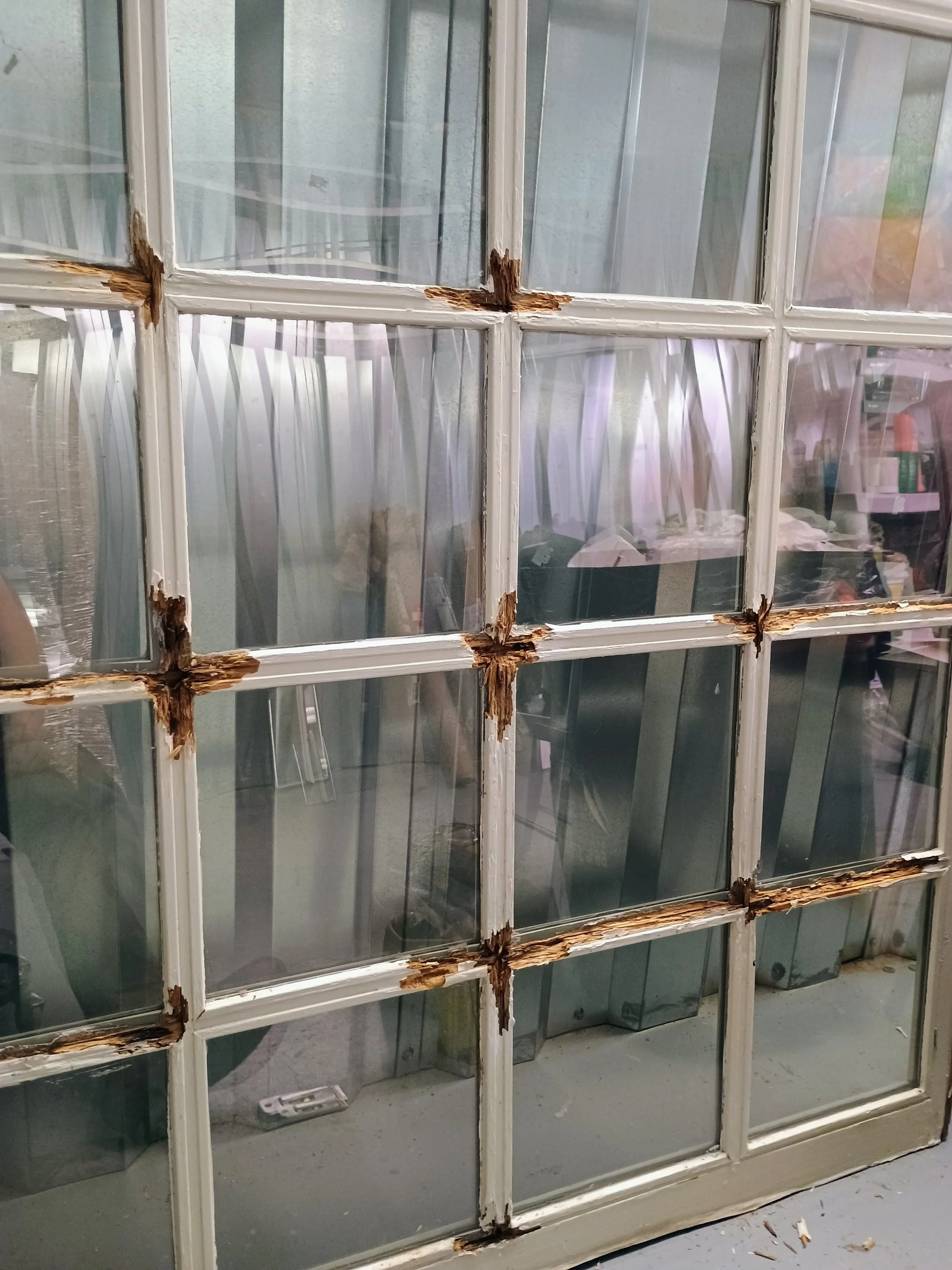 Close-up of a damaged white wooden window with multiple panes, showing peeling paint and rotting wood around the edges, with a cluttered storage area visible behind the window.