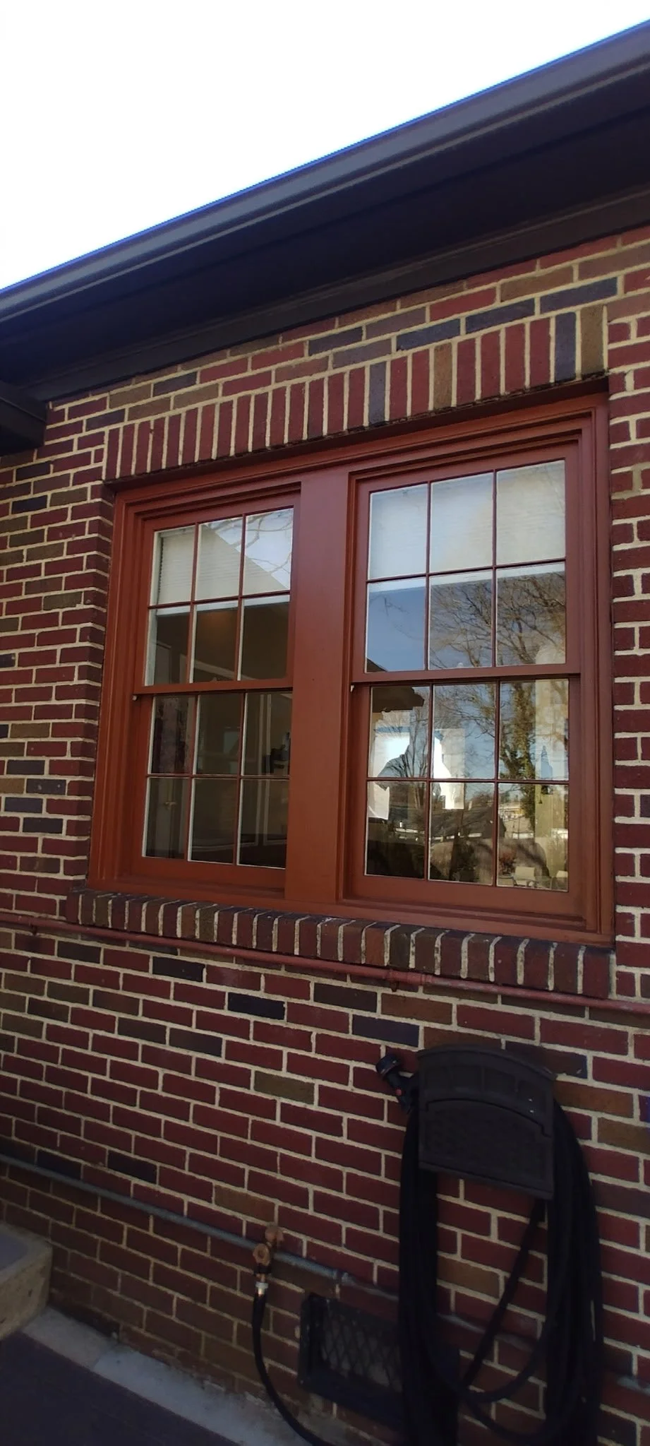 A large double-hung window with wooden trim on a brick house, reflecting trees and the sky.