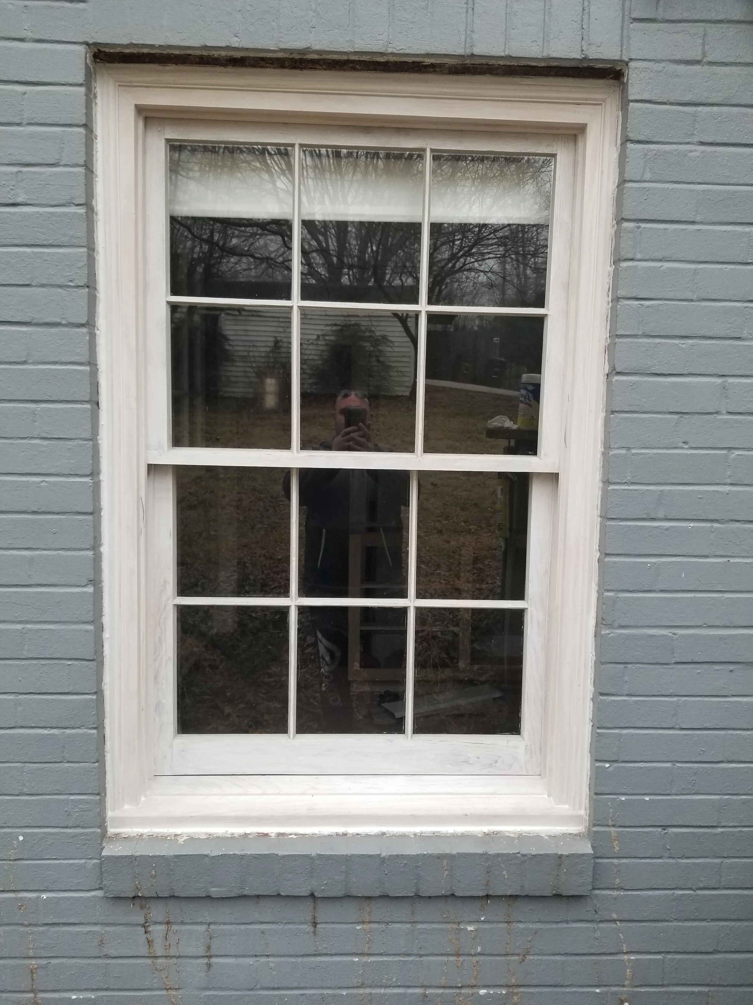 A window with white trim on a gray brick wall, reflecting the person taking the photo and the outdoor backyard with trees and a shed.