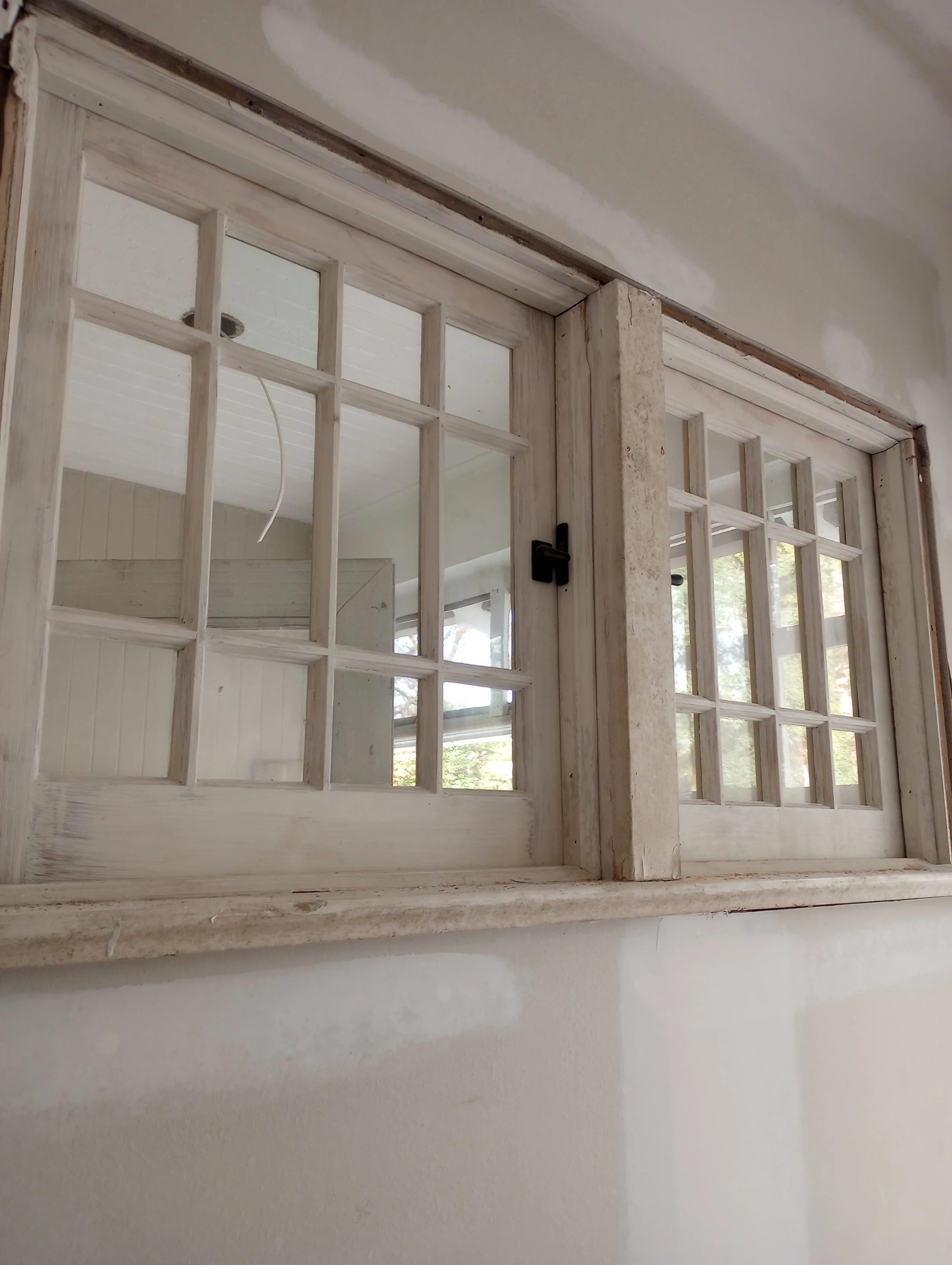 Close-up of a window with a grid of small panes, wood frame, and latch, looking into an interior room. The window appears to be newly installed or renovated with a light-colored distressed wood finish.