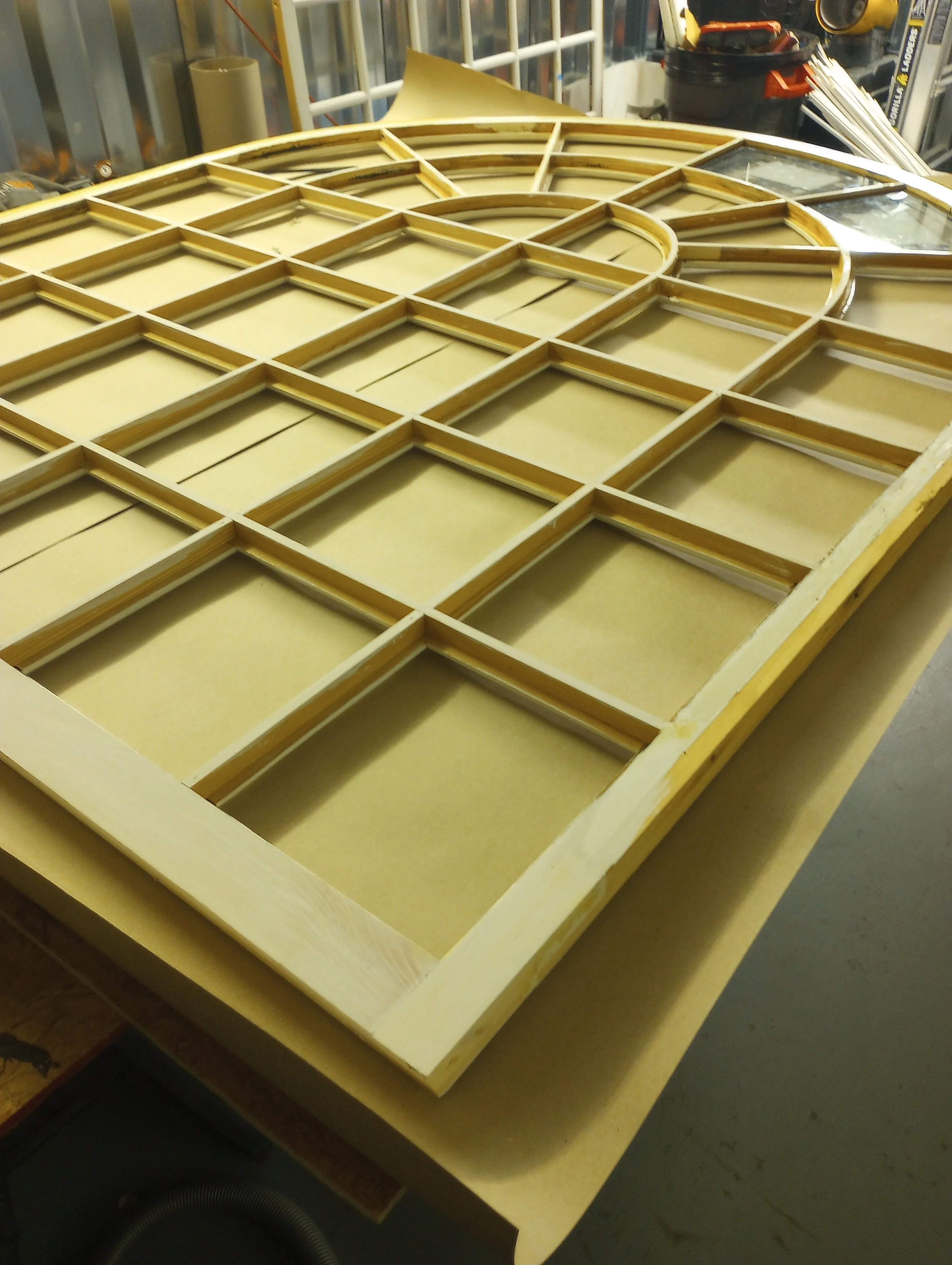 Wooden framework of a stained glass window on a workbench in a workshop, with tools and materials in the background.