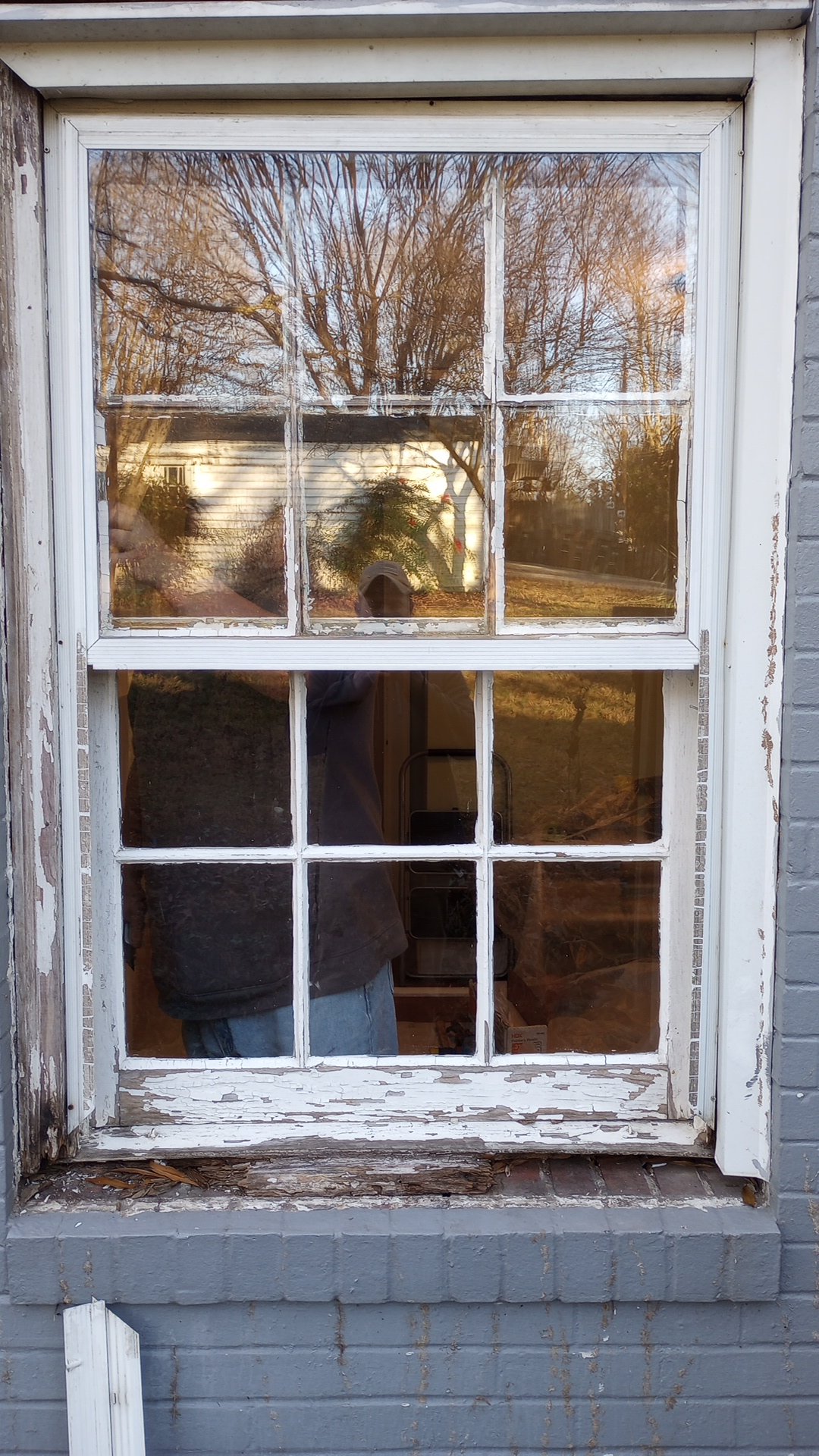 Refurbishing an old house window with peeling white paint and a brick exterior, showing a reflection of trees and a house.
